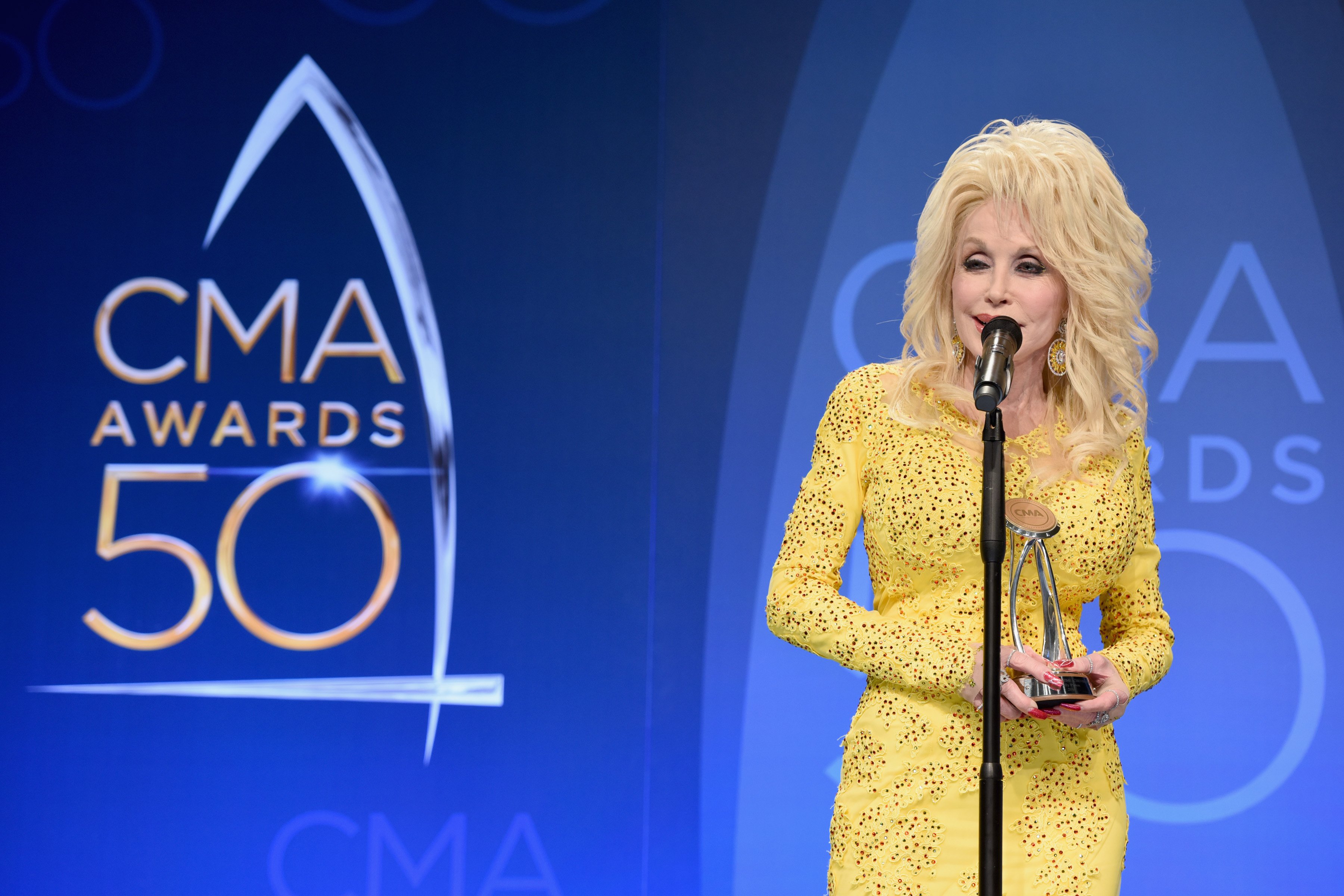 Dolly Parton at the 50th annual CMA Awards. She's speaking into a microphone on stage in a yellow dress.