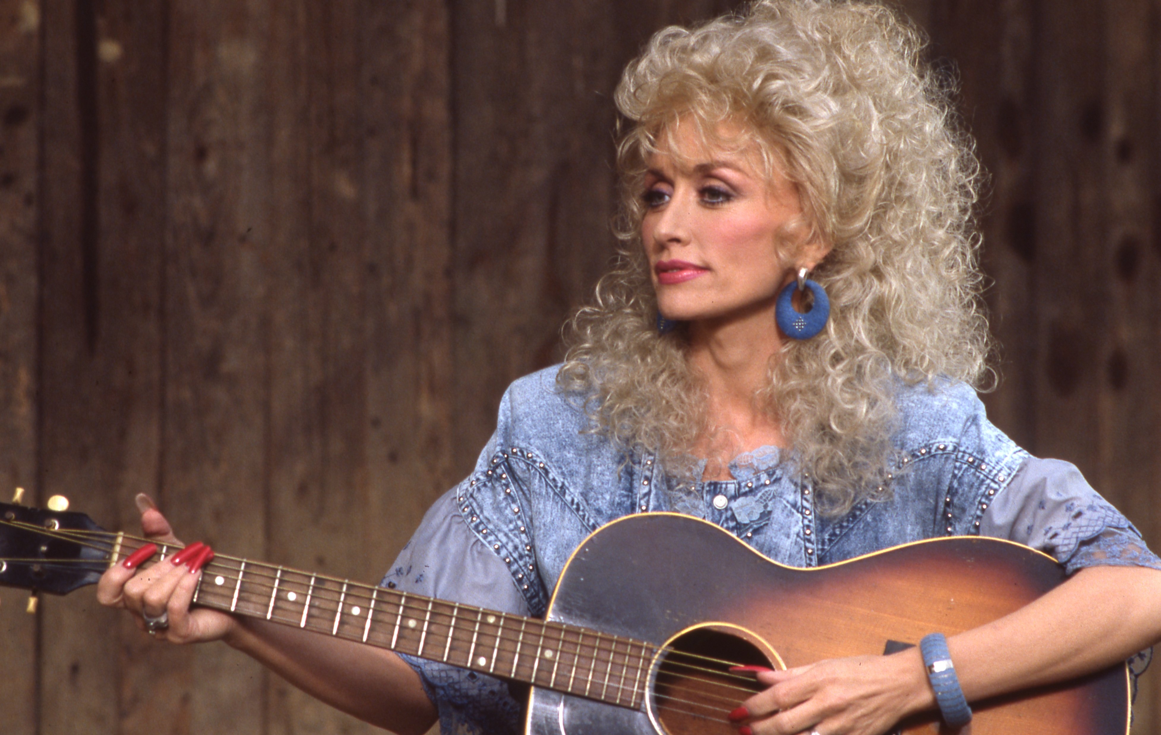 Dolly Parton playing the guitar. Her hair is big and curly. She's wearing a jean shirt, blue earrings, a blue bangle bracelet, and long, red nails.