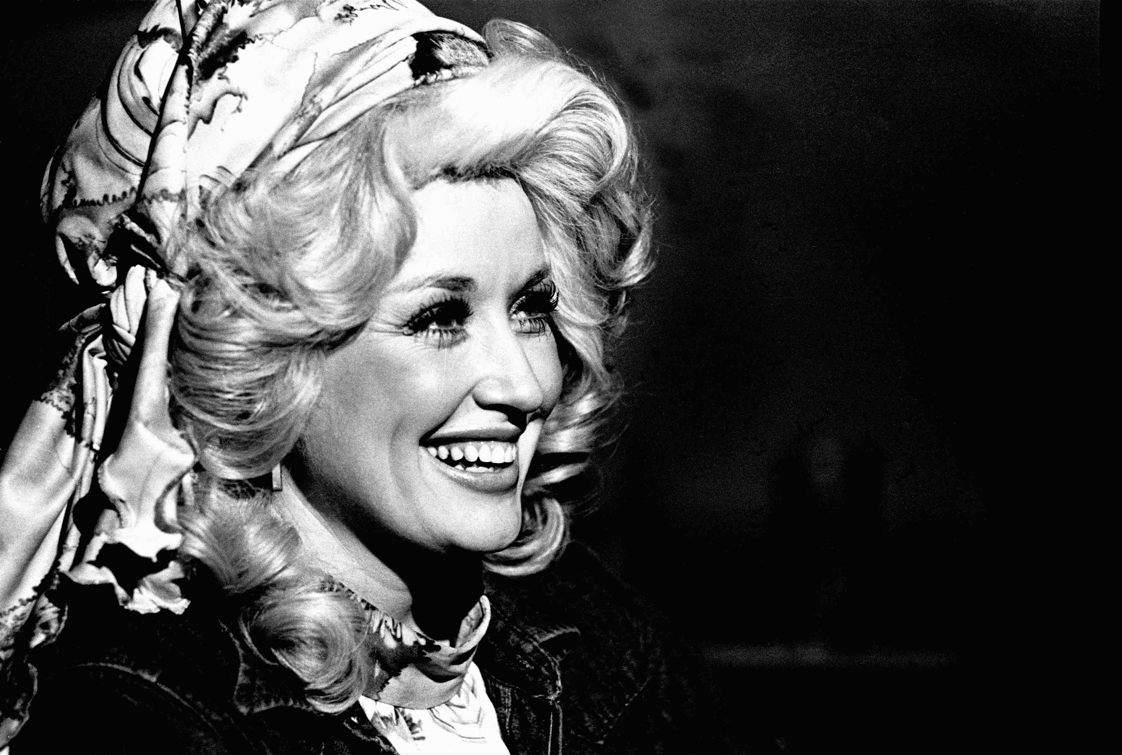 A close-up of Dolly Parton smiling her famous smile in black and white. Her hair is big and curly in a beehive with a scarf fastened around her head. The photo was taken in 1978.