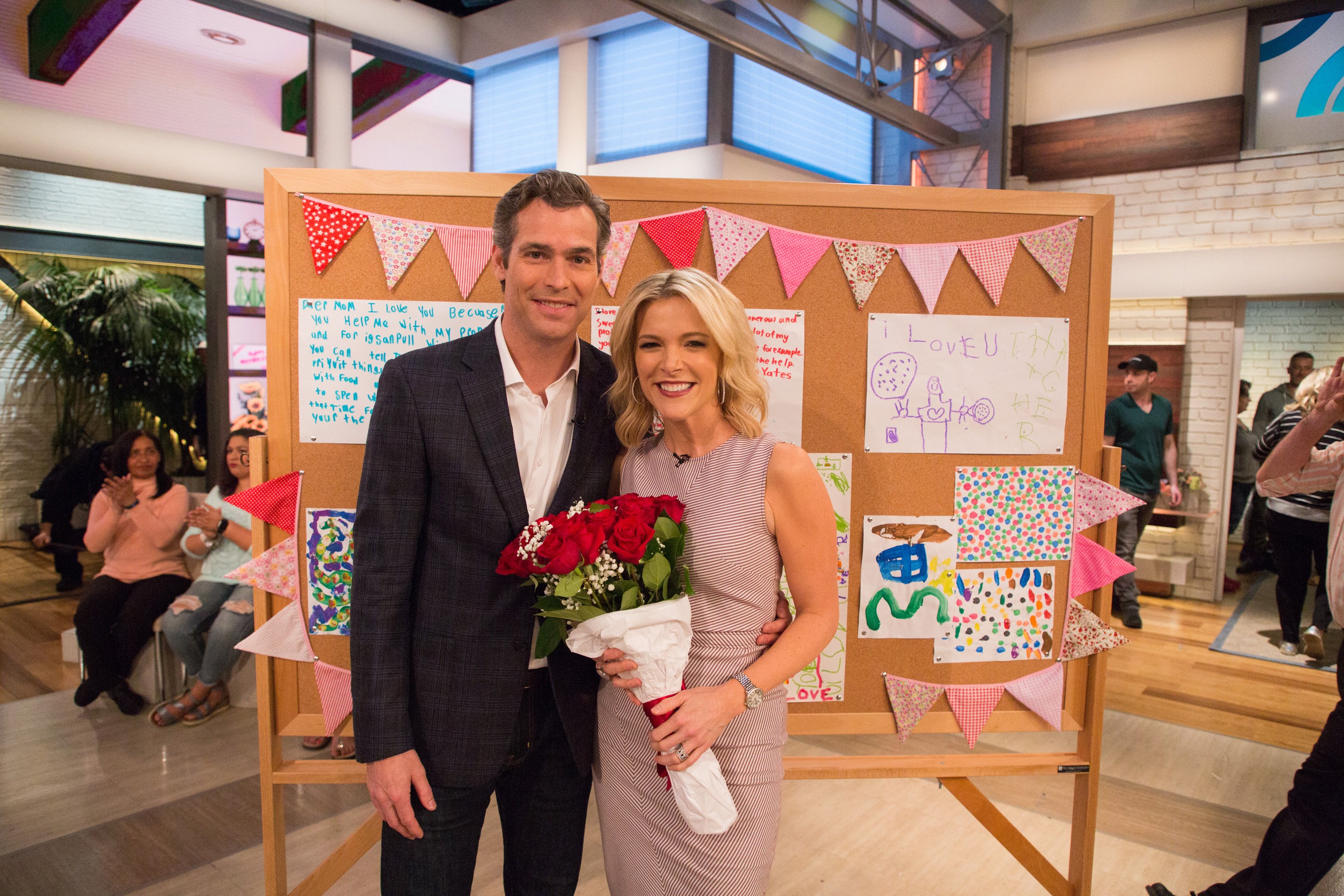 Doug Brunt in a dark jacket standing with wife Megyn Kelly in a gray dress holding a bouquet of flowers