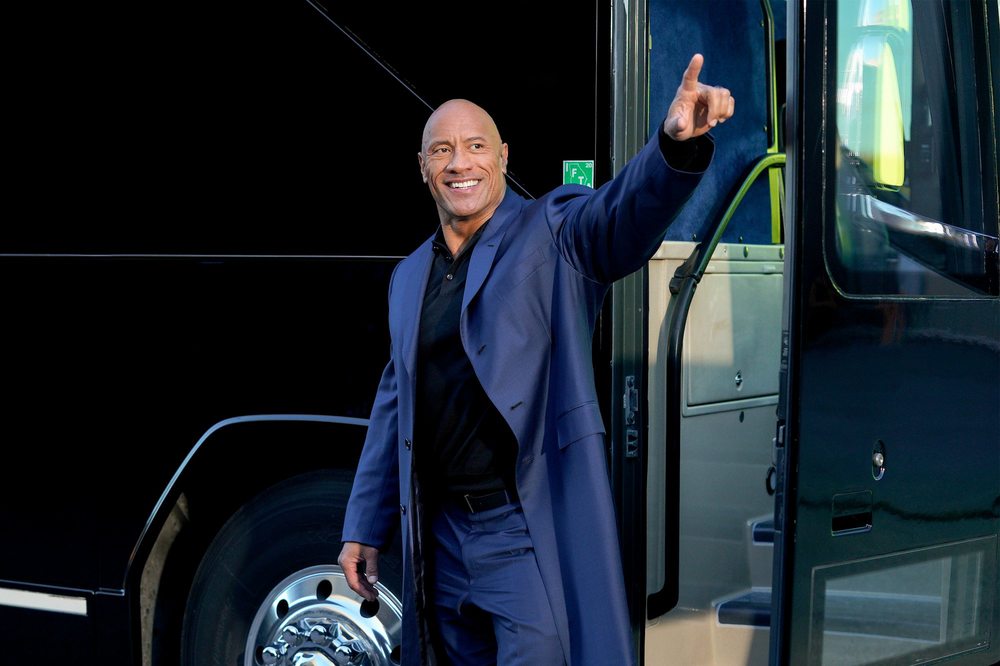 Dwayne Johnson smiling and pointing, standing in front of a van