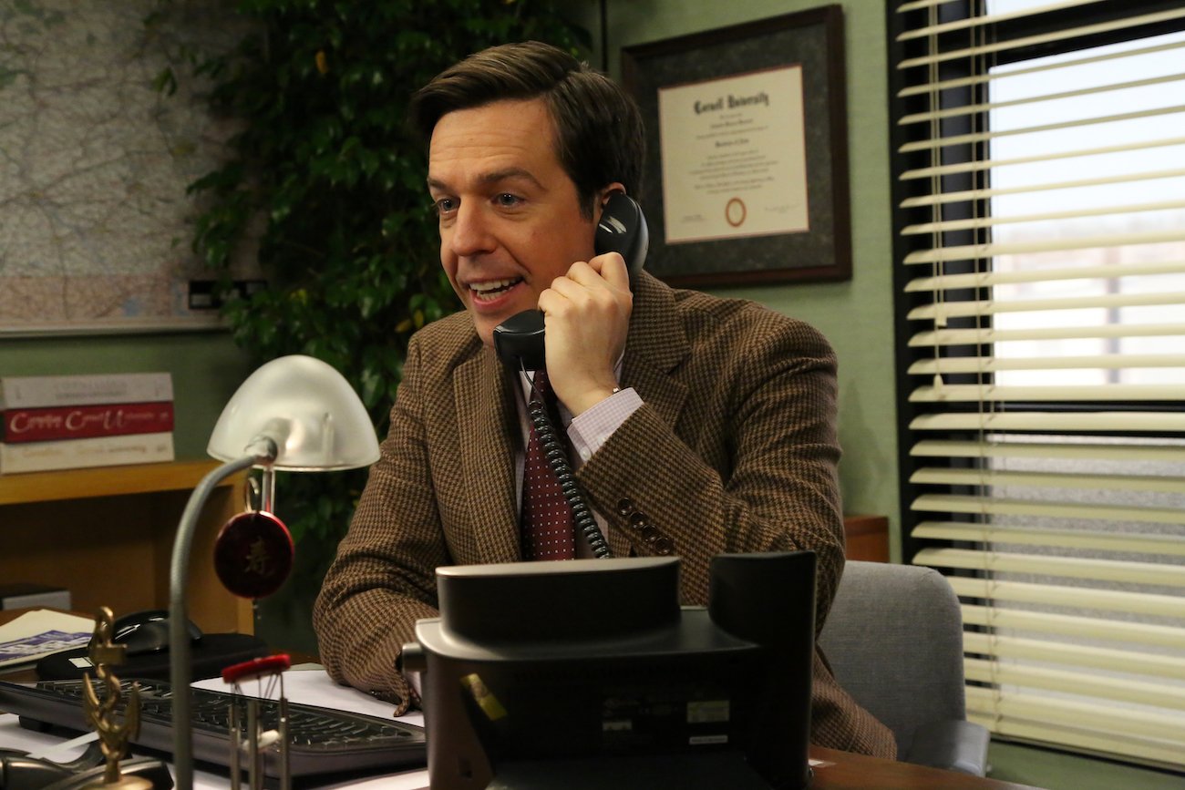 Ed Helms sits at a desk talking on the phone in a brown jacket and maroon tie in a scene from 'The Office'