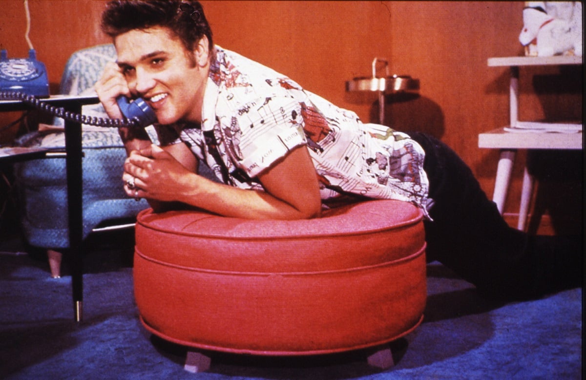 Elvis Presley in a posed color photo, leaning on a stool as he talks on the phone