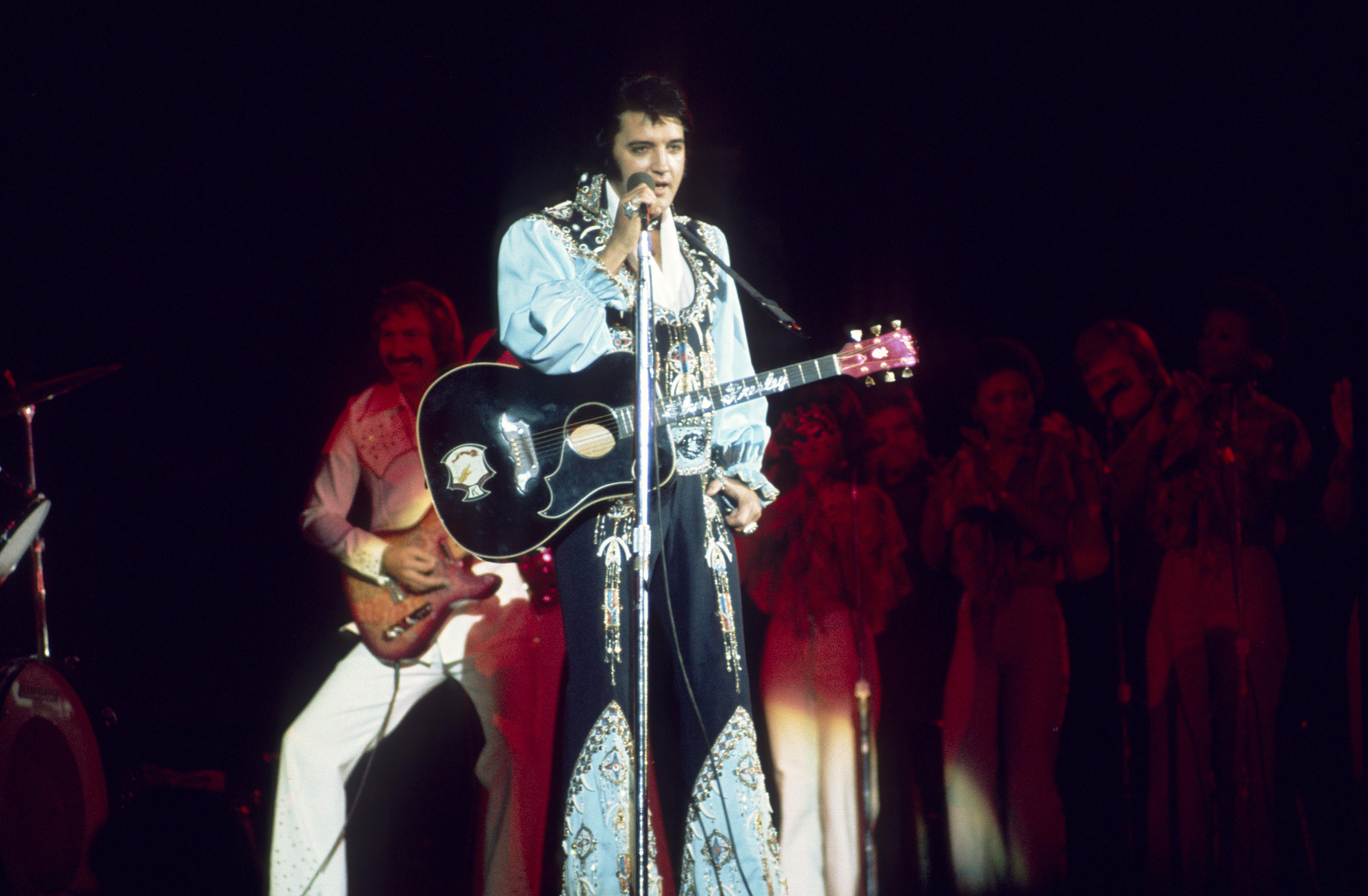 Elvis Presley performing on stage. He's singing into a stand microphone and holding a black guitar.