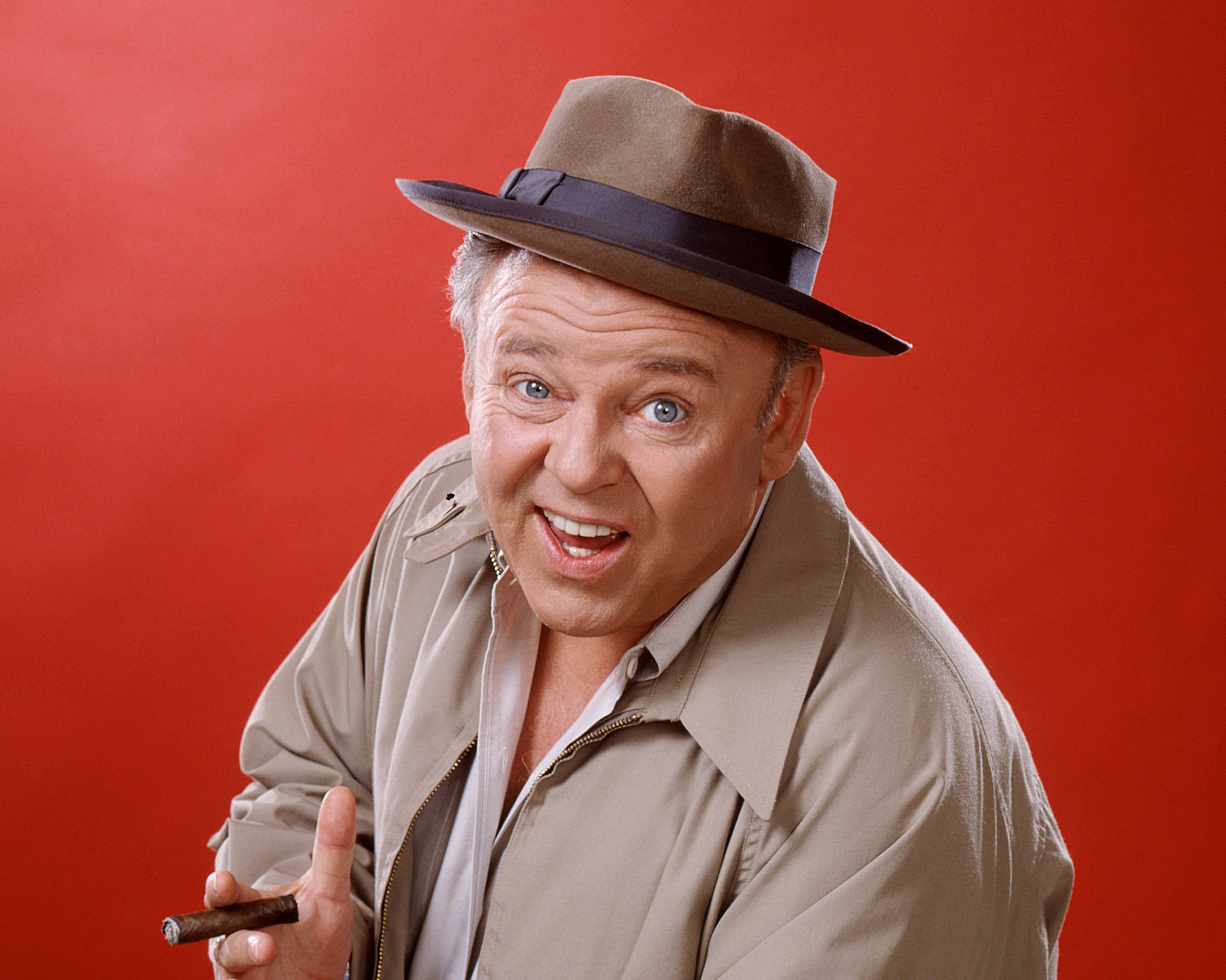Carroll O'Connor, wearing a brown hat with a dark blue band and a beige jacket, and holding a cigar against a red background as Archie Bunker in 'All in the Family', circa 1975
