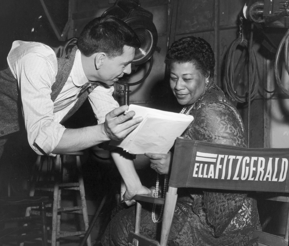 American actor and director Jack Webb discusses a script with American jazz singer Ella Fitzgerald behind the scenes, possibly on the set of Webb's film, 'Pete Kelly's Blues,' in which Fitzgerald had a role, 1955