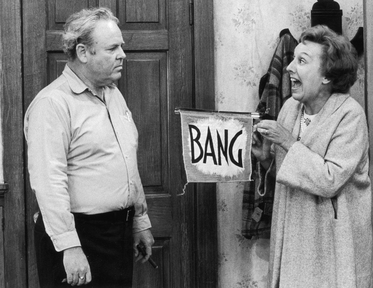 Carroll O'Connor, left, watches as Jean Stapleton points a toy gun with a flag that reads, 'Bang,' 1975