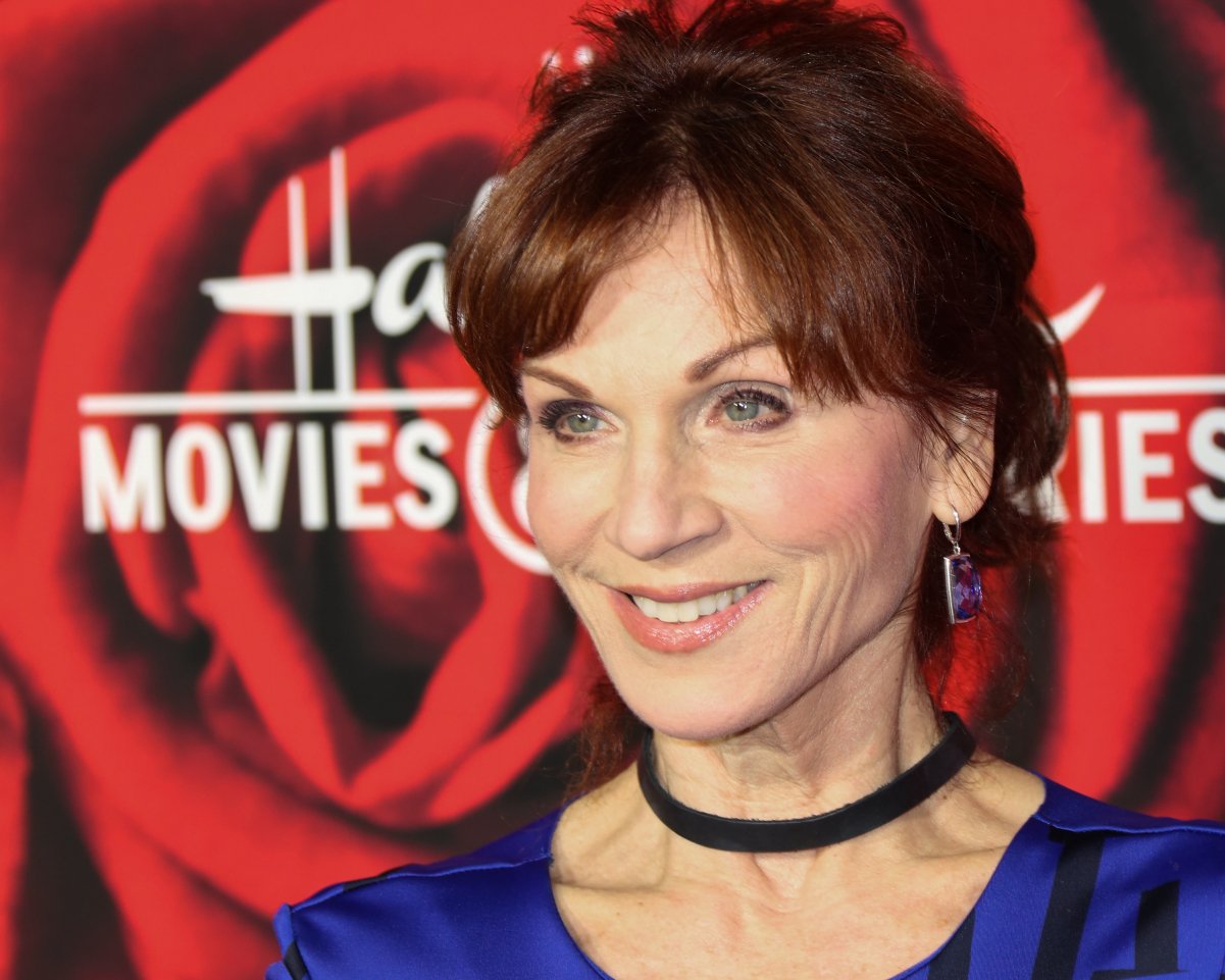 Actor Marilu Henner attends a Hallmark Channel and Hallmark Movies and Mysteries event in 2017. She wears a black choker and blue blouse with her red hair in a bun.