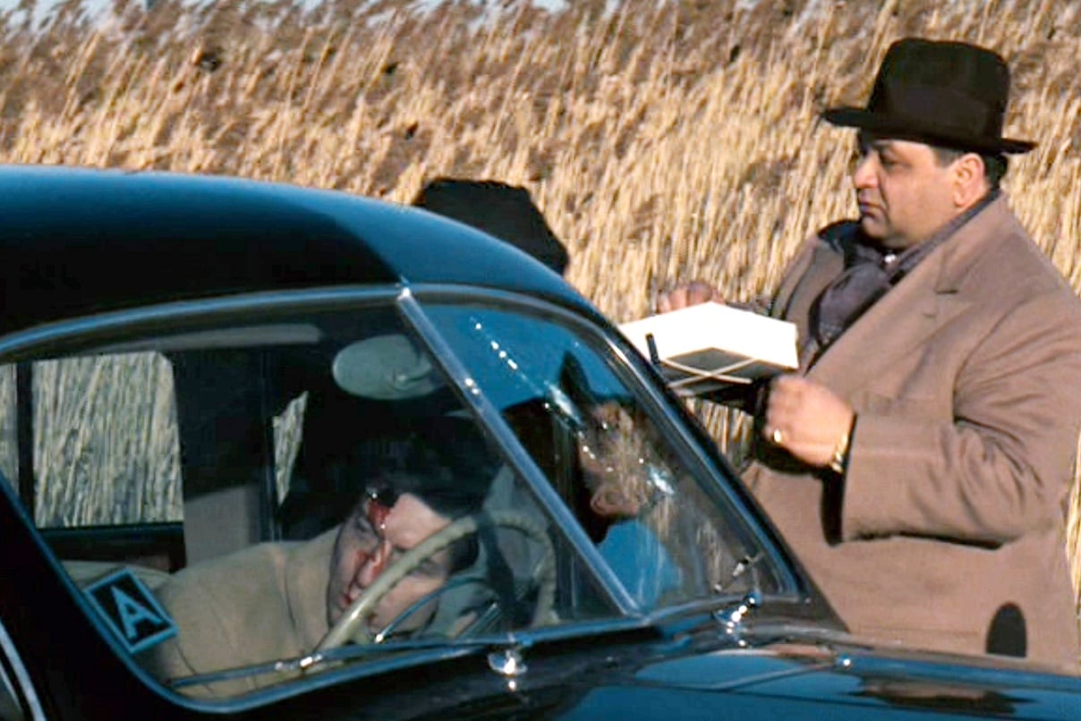 Richard Castellano stands next to a car holding a box of cannoli during a scene from 'The Godfather.' The car's driver is bloody and dead