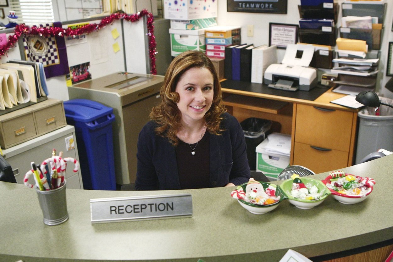 The Office star Jenna Fischer as Pam Beesly sits at her desk at reception and smiles at the camera