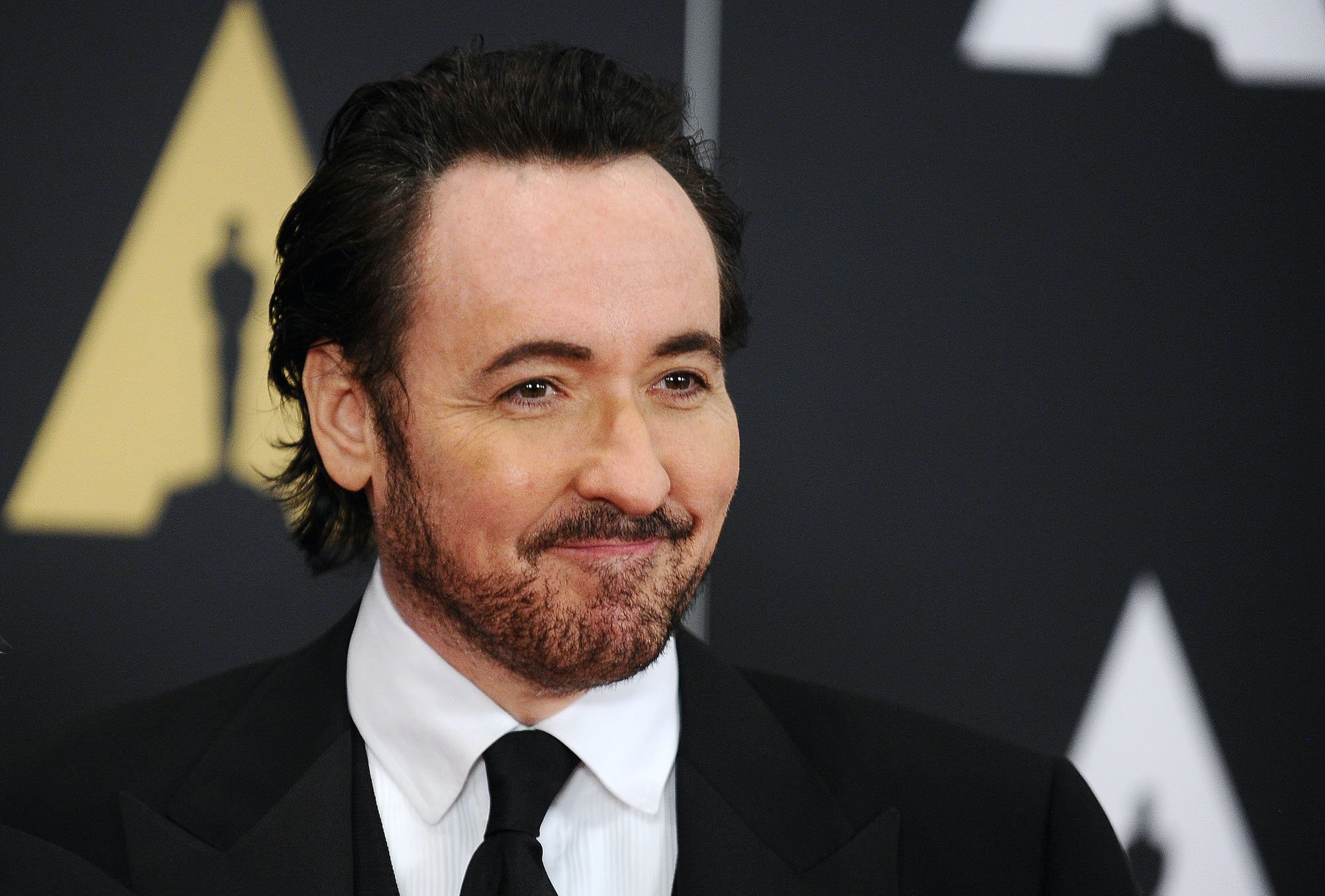 John Cusack smiling in front of a black background
