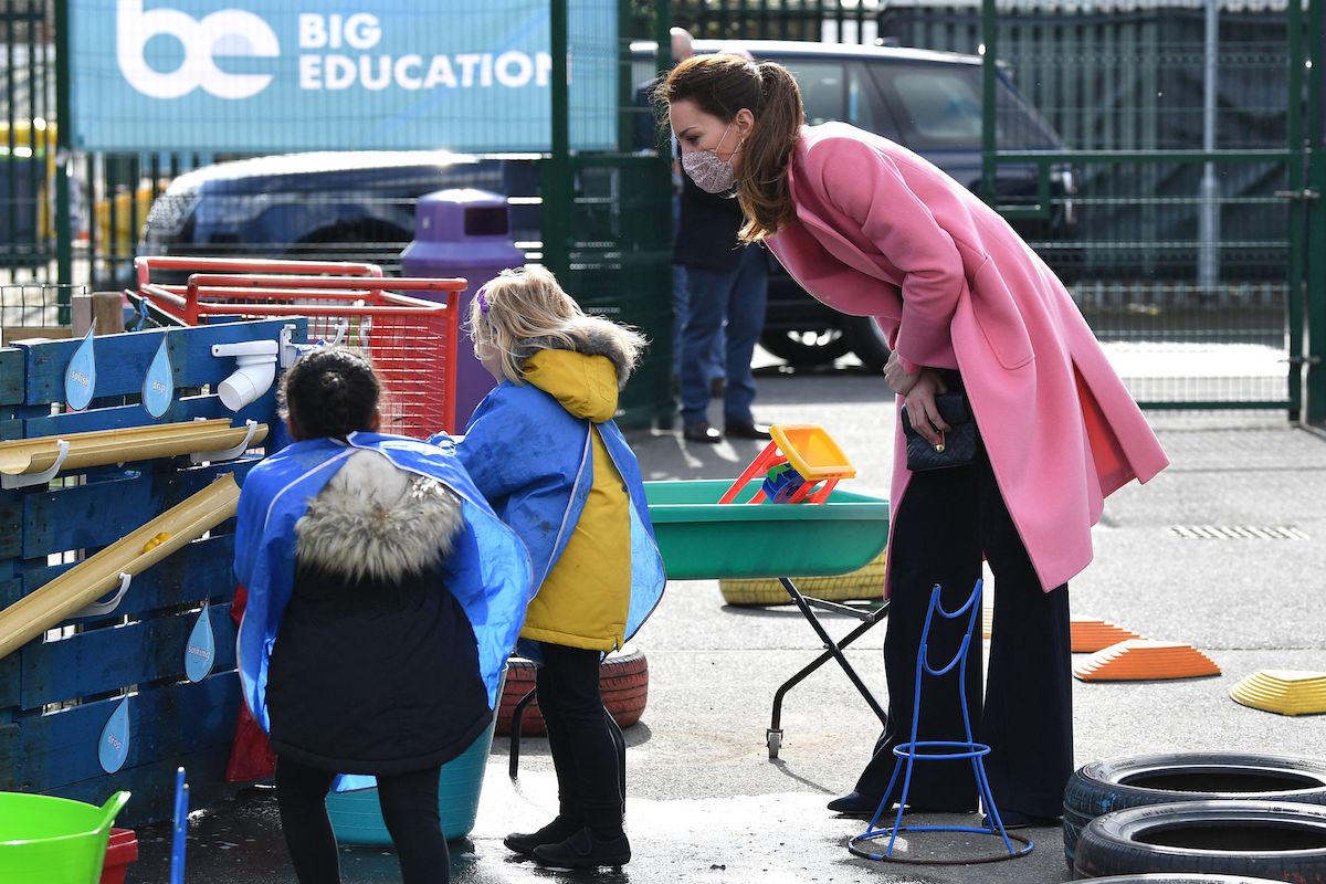 Kate Middleton leaning over and talking to children wearing a pink peacoat and black pants.