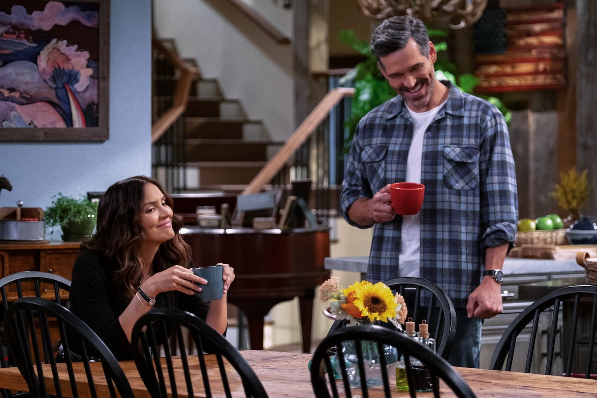 Katharine McPhee sits at a table holding a coffee cup while Eddie Cibrian stands next to her holding a coffee cup during a scene from 'Country Comfort'