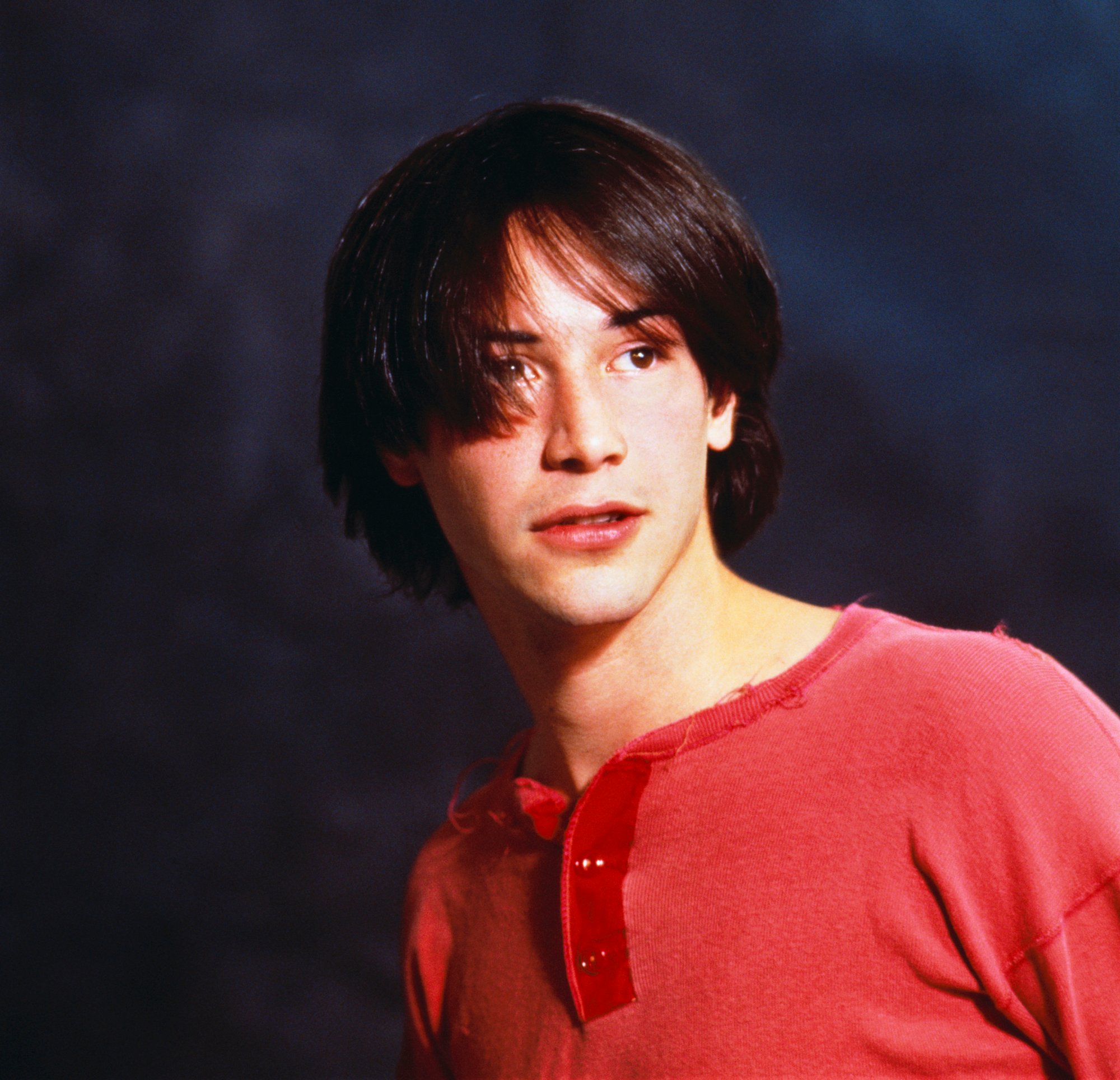 A young Keanu Reeves, with a wispy bowl haircut and a red henley shirt, poses in a photo studio