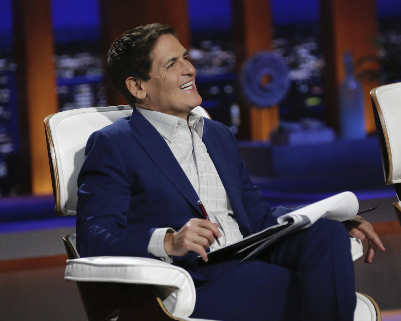 Mark Cuban sitting on the panel of of 'Shark Tank' smiling in a dark jacket and white shirt