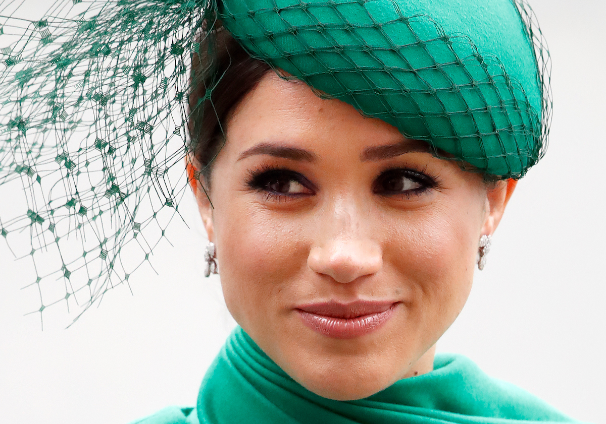 Meghan, Duchess of Sussex is smirking while wearing a green dress and matching hat.