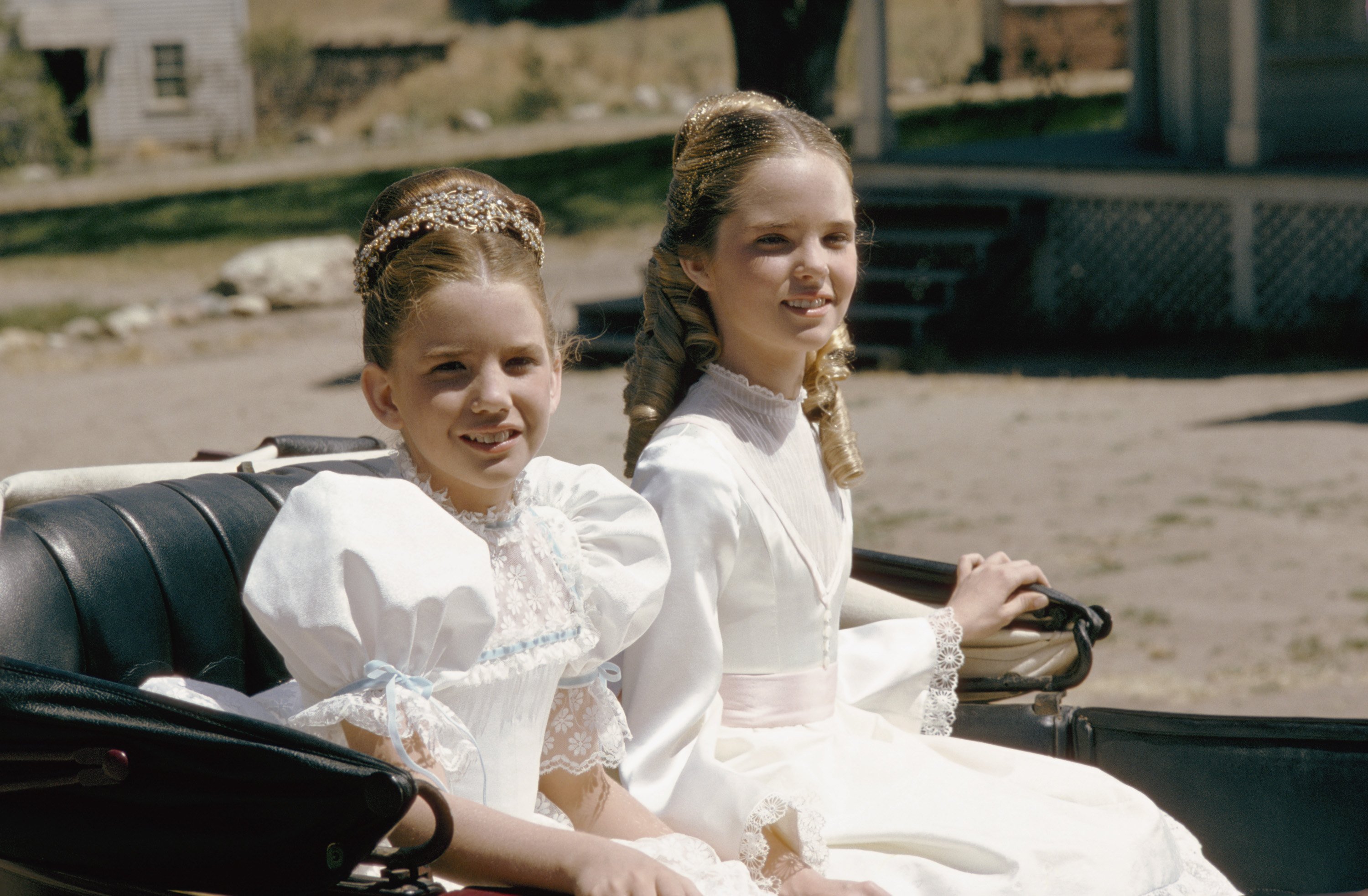 Melissa Gilbert and Melissa Sue Anderson sit in a carriage on the set of 'Little House on the Prairie.' They are in white dresses.