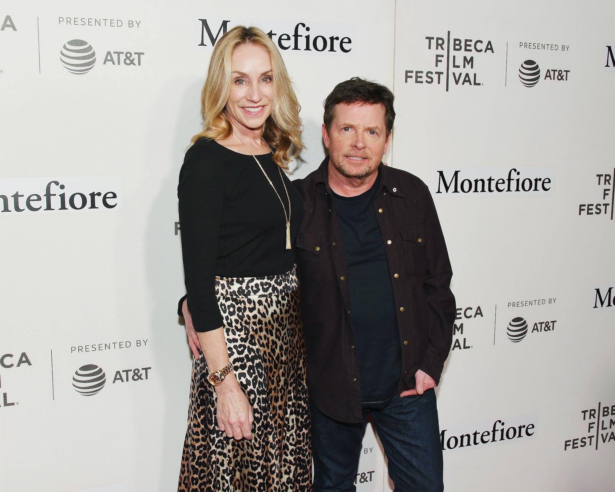 (L-R) Tracy Pollan and Michael J. Fox smiling in front of a white background with repeating logos