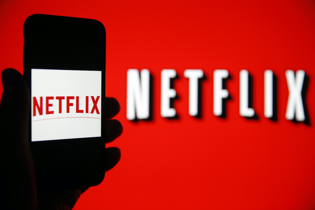 Netflix logo is seen on the screen of an iPhone in front of a computer screen showing a Netflix logo