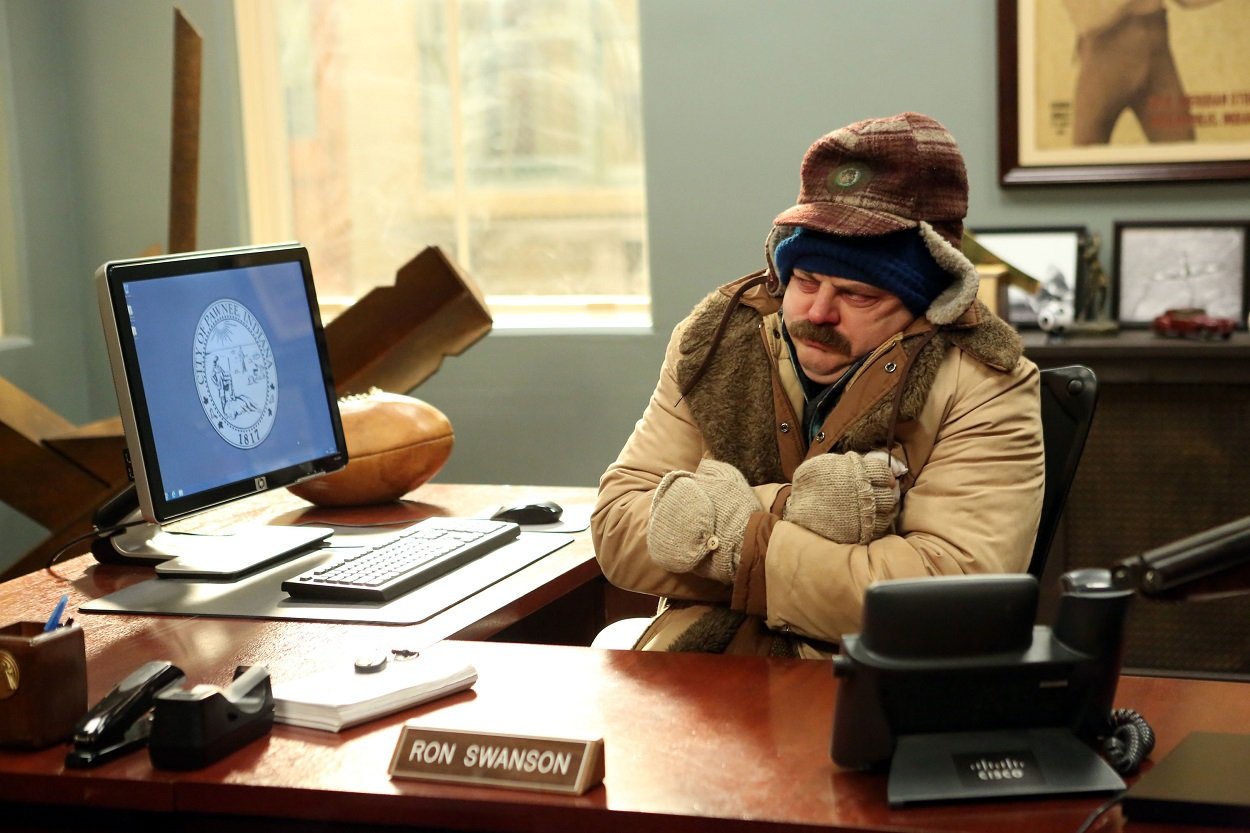 Parks and Recreation cast member Nick Offerman as Ron Swanson at his desk bundled up in a coat