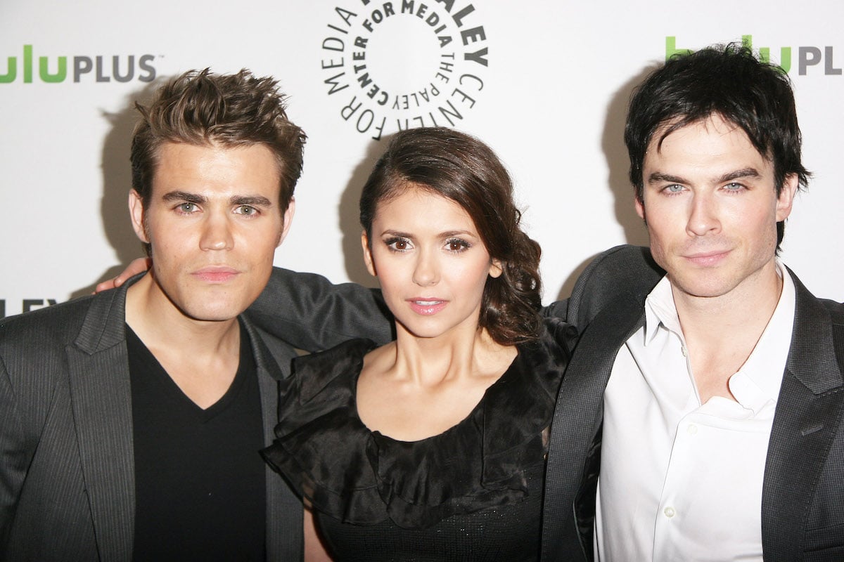 Paul Wesley, Nina Dobrev, and Ian Somerhalder wearing black and white and standing in front of a white and green backdrop | Tommaso Boddi/WireImage