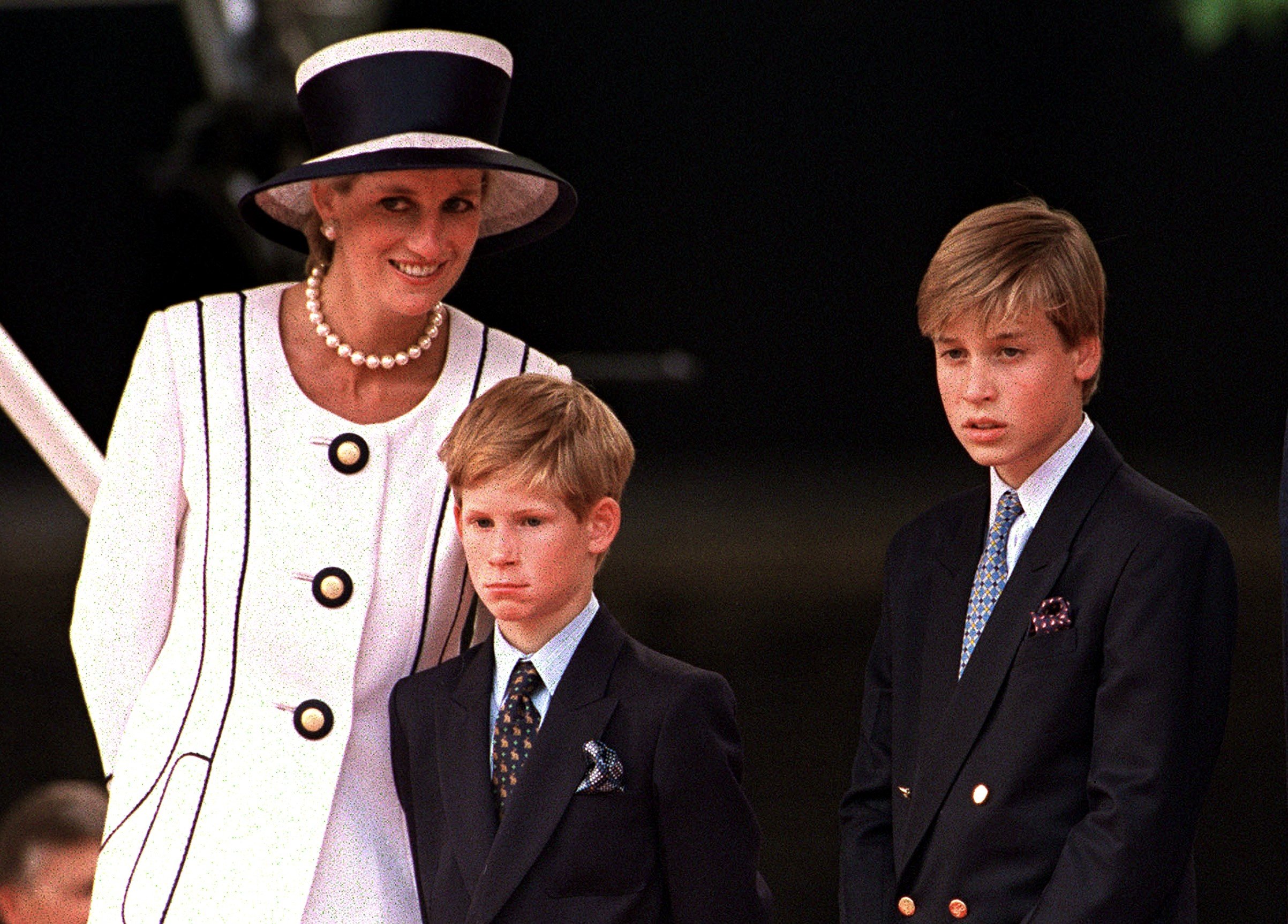 Prince Harry and Prince William dressed in suits next to Princess Diana wearing a white-and-black outfit with a fascinator at a parade on V.J. Day in 1994