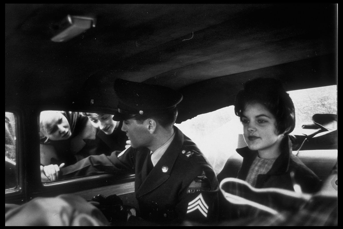 Elvis Presley wearing his military uniform while chatting with fans through the window of a car. A young Priscilla Beaulieu, then his girlfriend, is seated next to him.