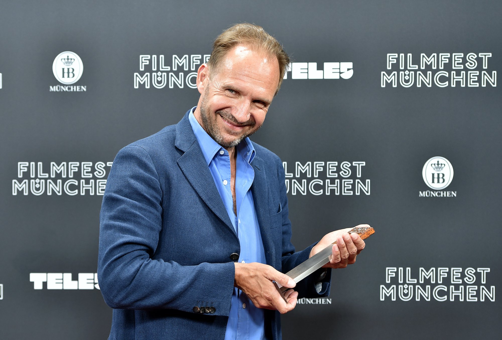 Ralph Fiennes smiling in front of a gray background, holding an award