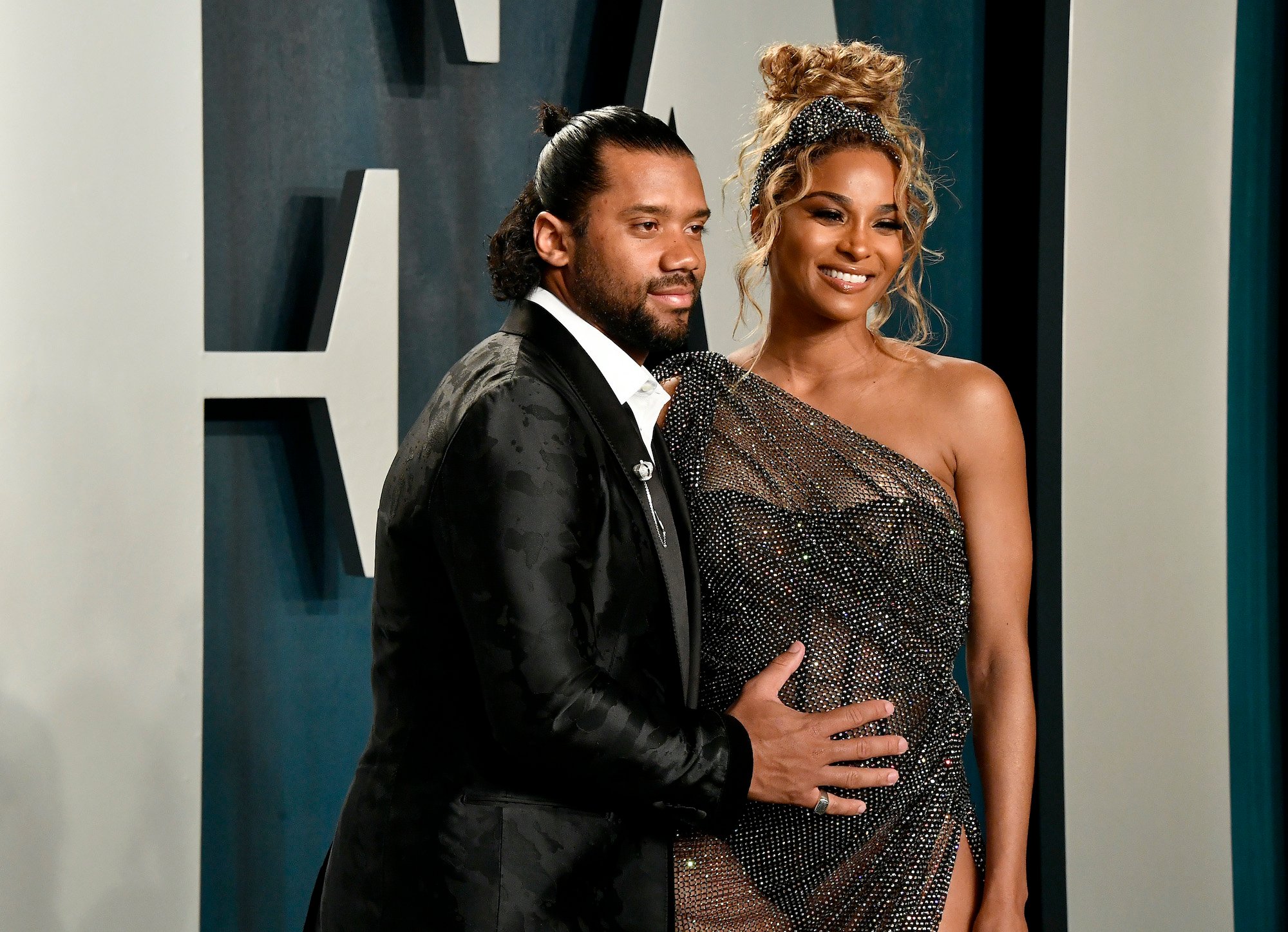 (L-R) Russell Wilson and Ciara smiling in front of a blue background