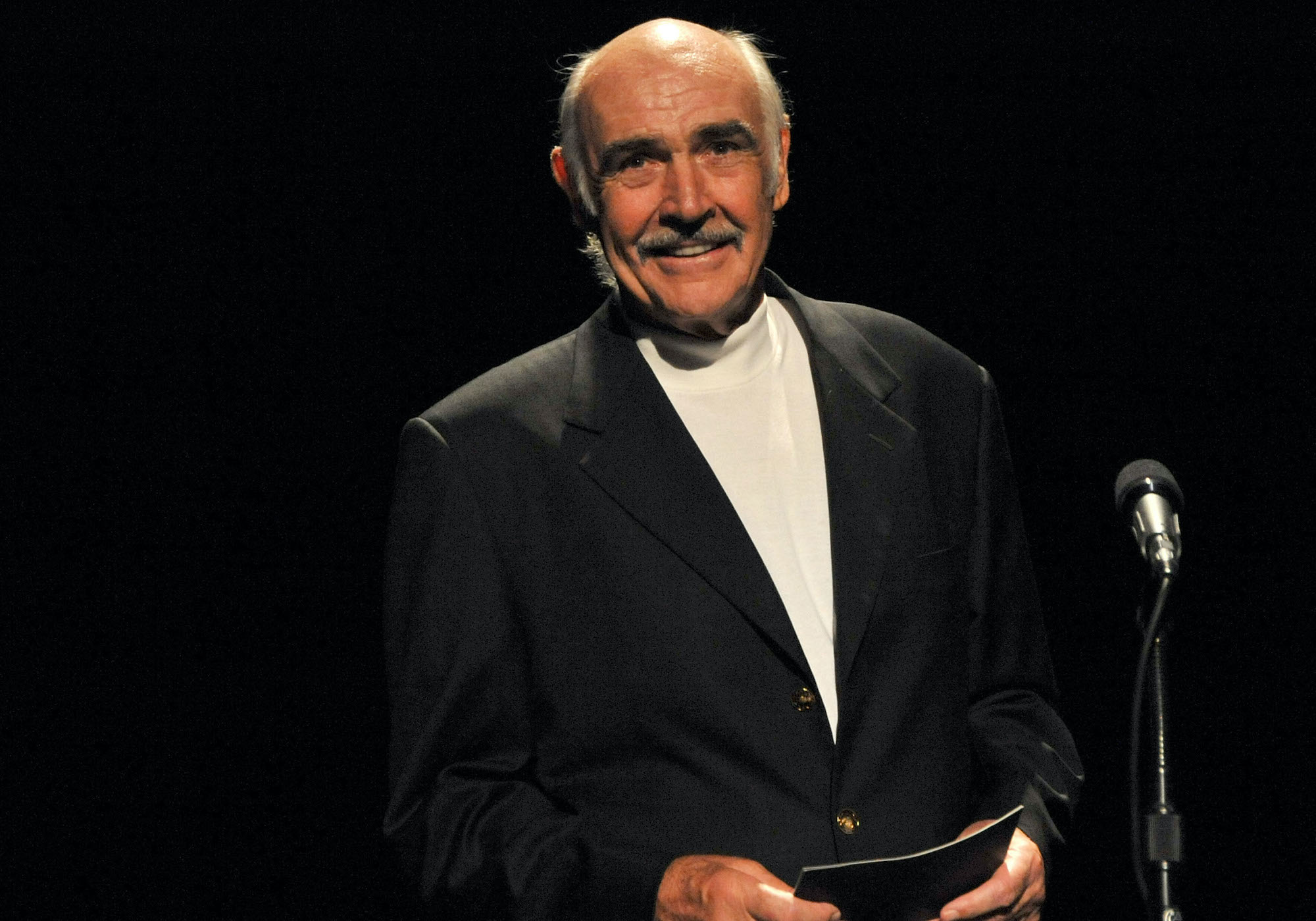 Sean Connery smiling behind a microphone in front of a black background