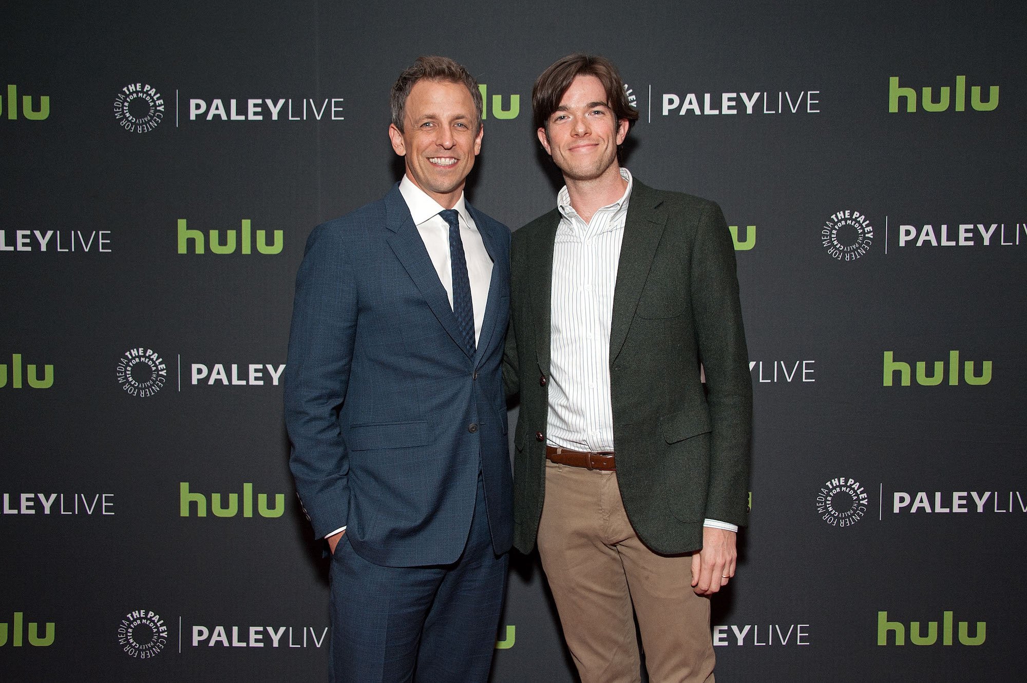 (L-R) Seth Meyers and John Mulaney smiling in front of a black background