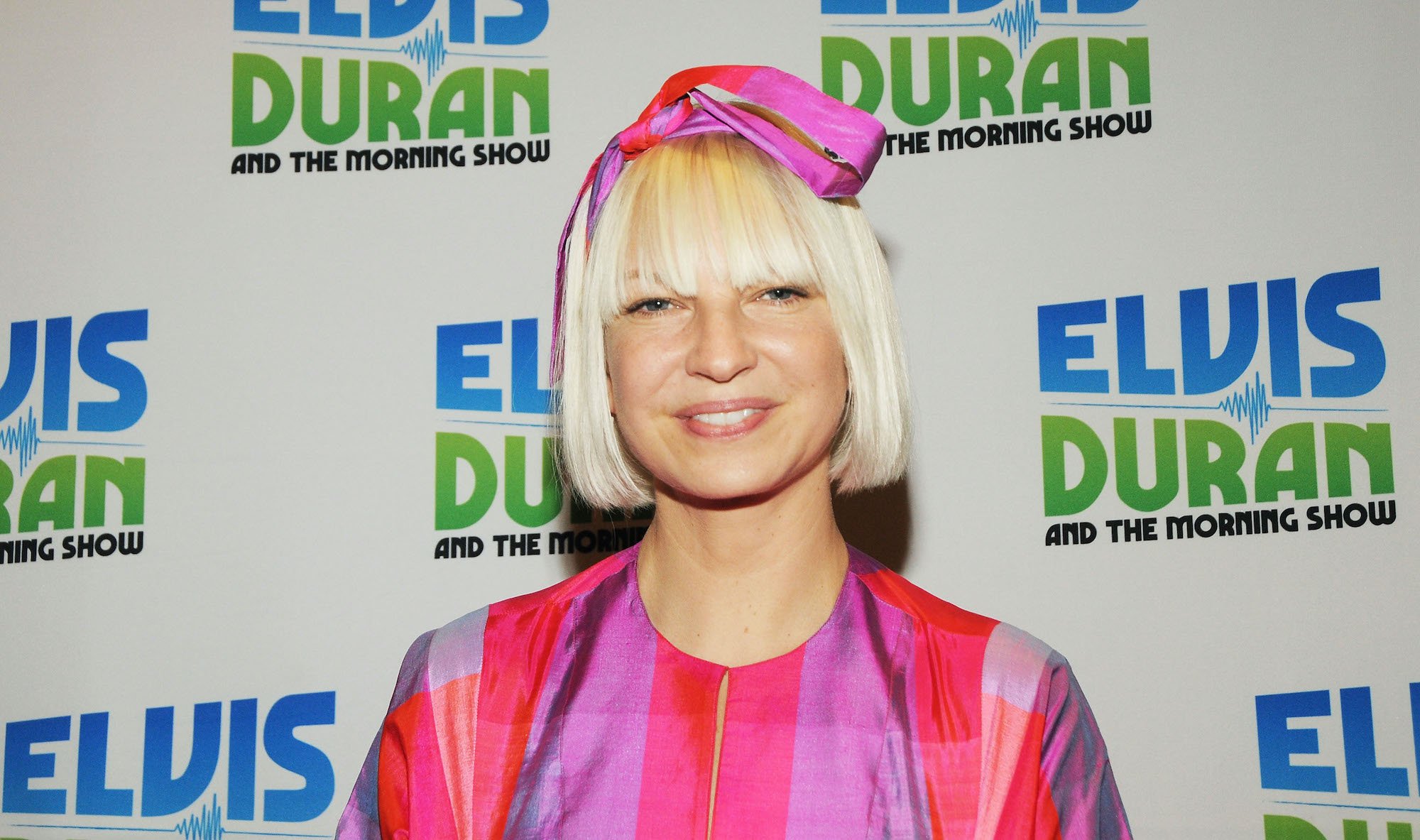 Sia smiling in front of a white background with repeating logos