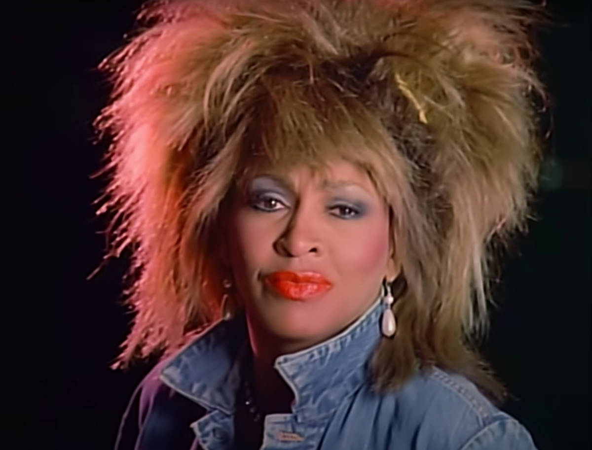 Tina Turner with her hair teased high wearing a blue denim jacket and bright red lipstick in the 'What's Love Got to Do With It' music video
