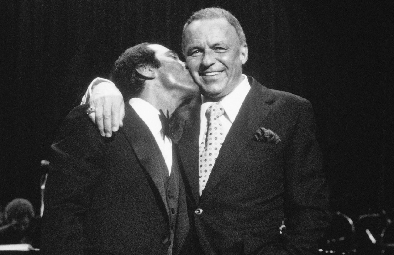 Frank Sinatra smiles and holds his arm around Paul Anka, who kisses him on the cheek