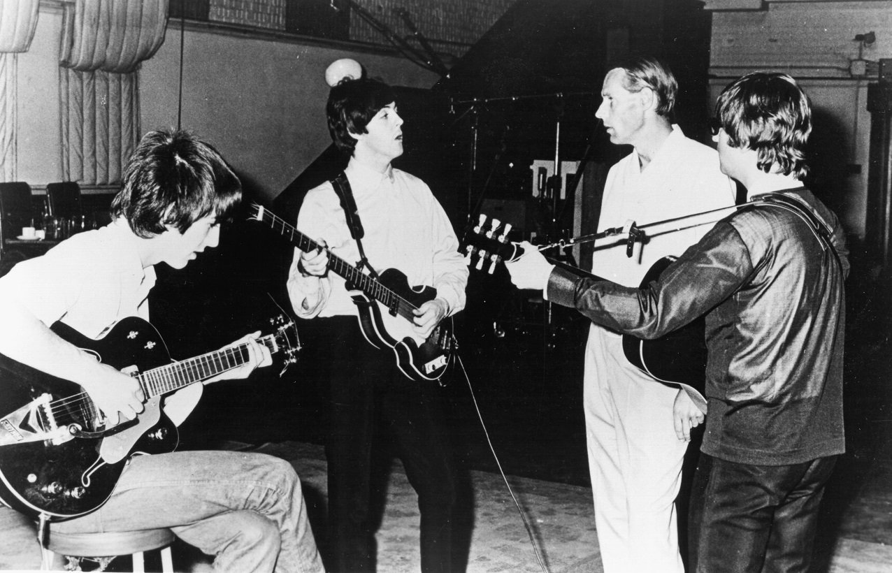 Paul McCartney, standing and holding a bass guitar, looks at George Martin as a seated George Harrison and standing John Lennon play guitar