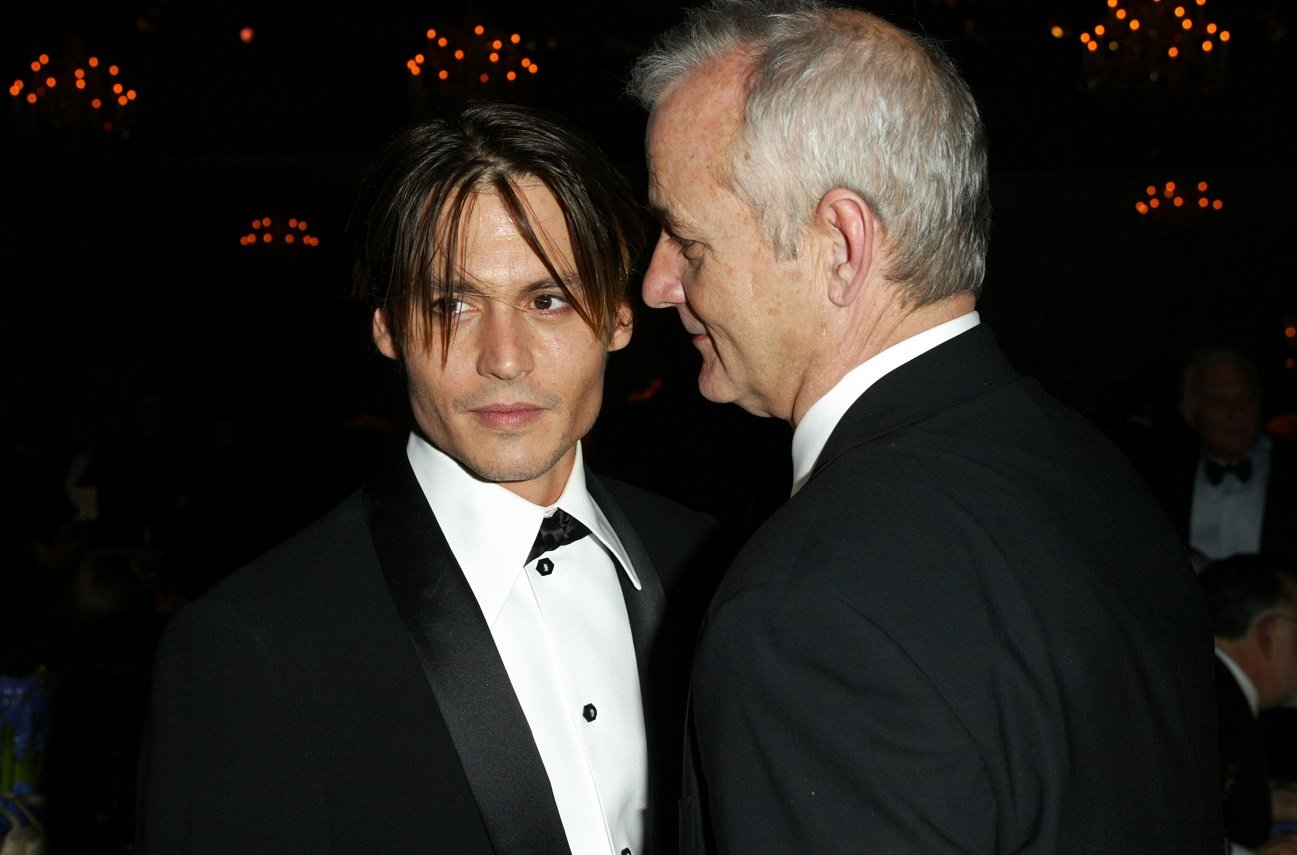 Johnny Depp has a slight smile and looks away as Bill Murray speaks very closely to his left ear