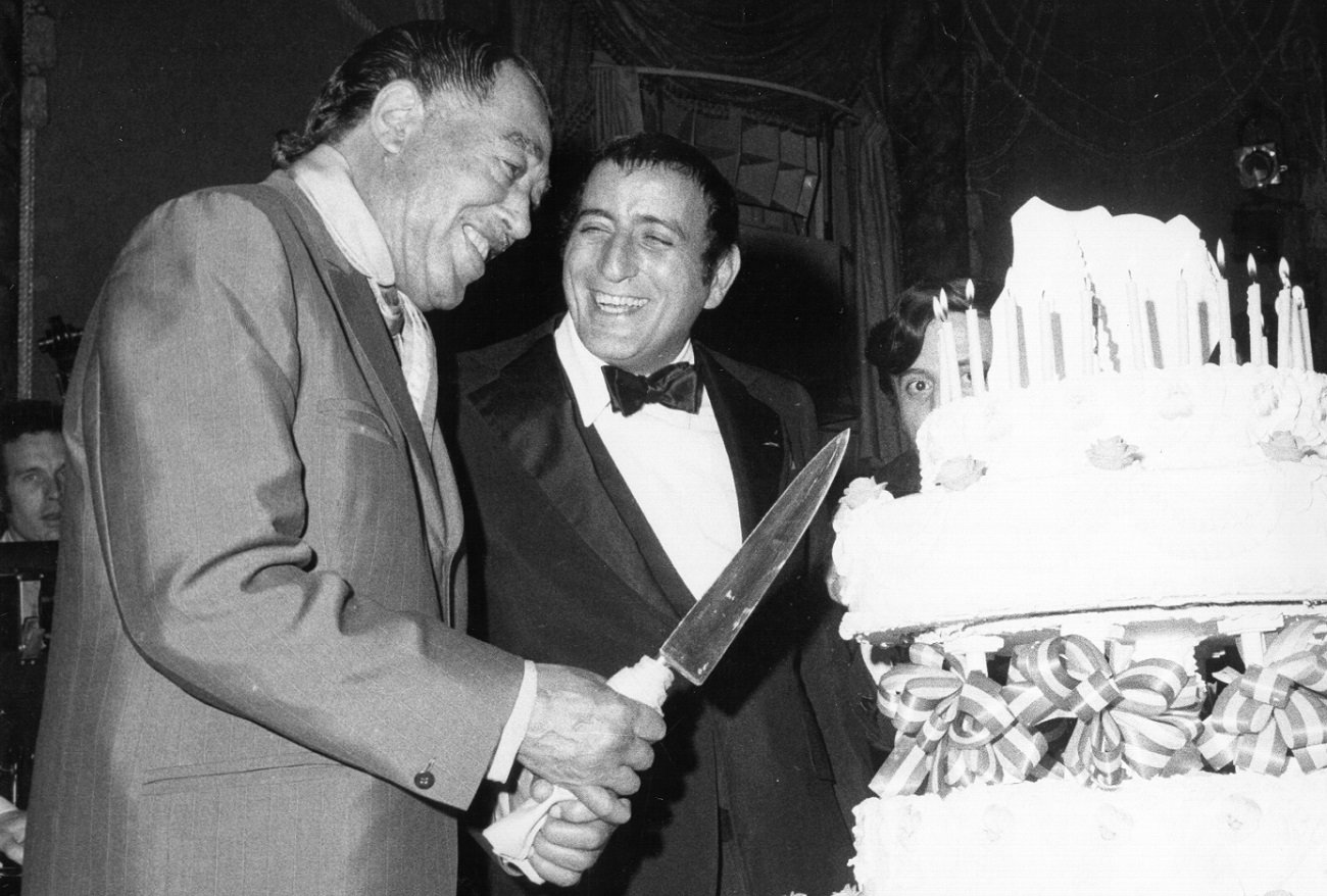 Duke Ellington holding a knife to cut a cake while Tony Bennett smiles next to him