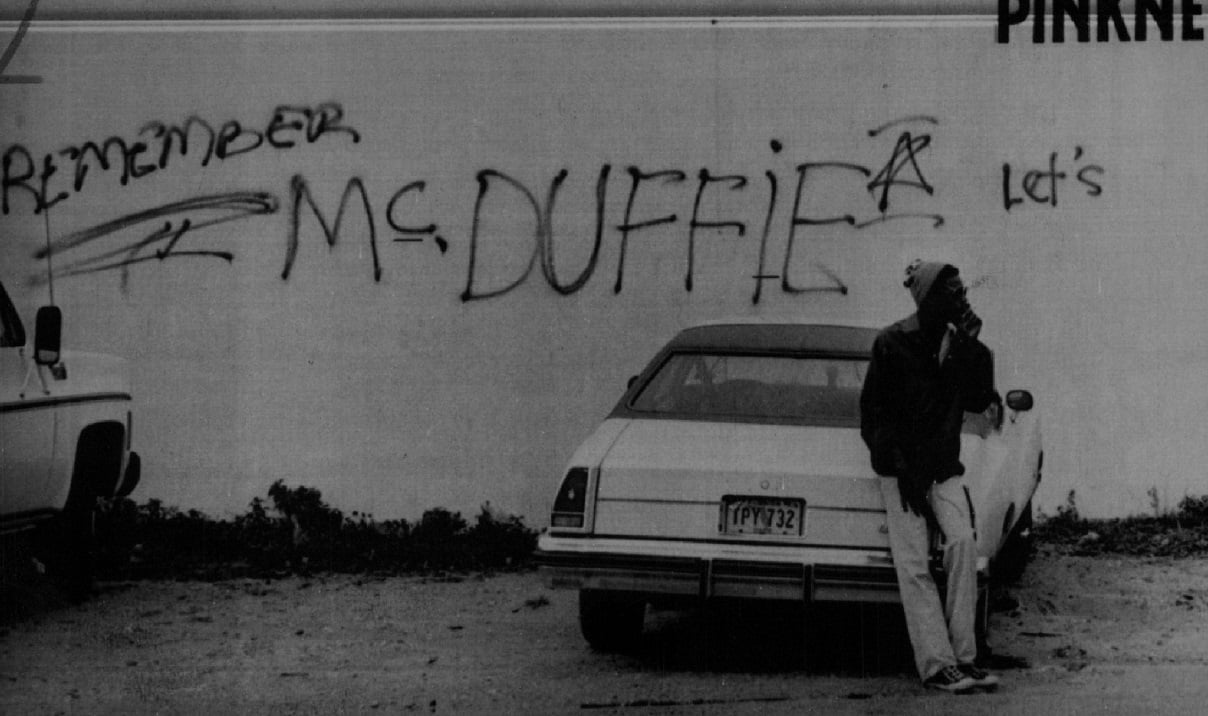 A young Black man stands in front of a graffiti-covered wall in Miami, a reminder of the riots set off when policemen were acquitted for the murder of Arthur McDuffie