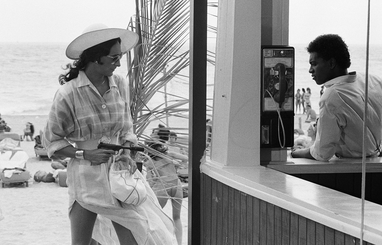 A man in drag holding a gun approaches a man standing at a beachfront cafe in the 'Miami Vice' pilot