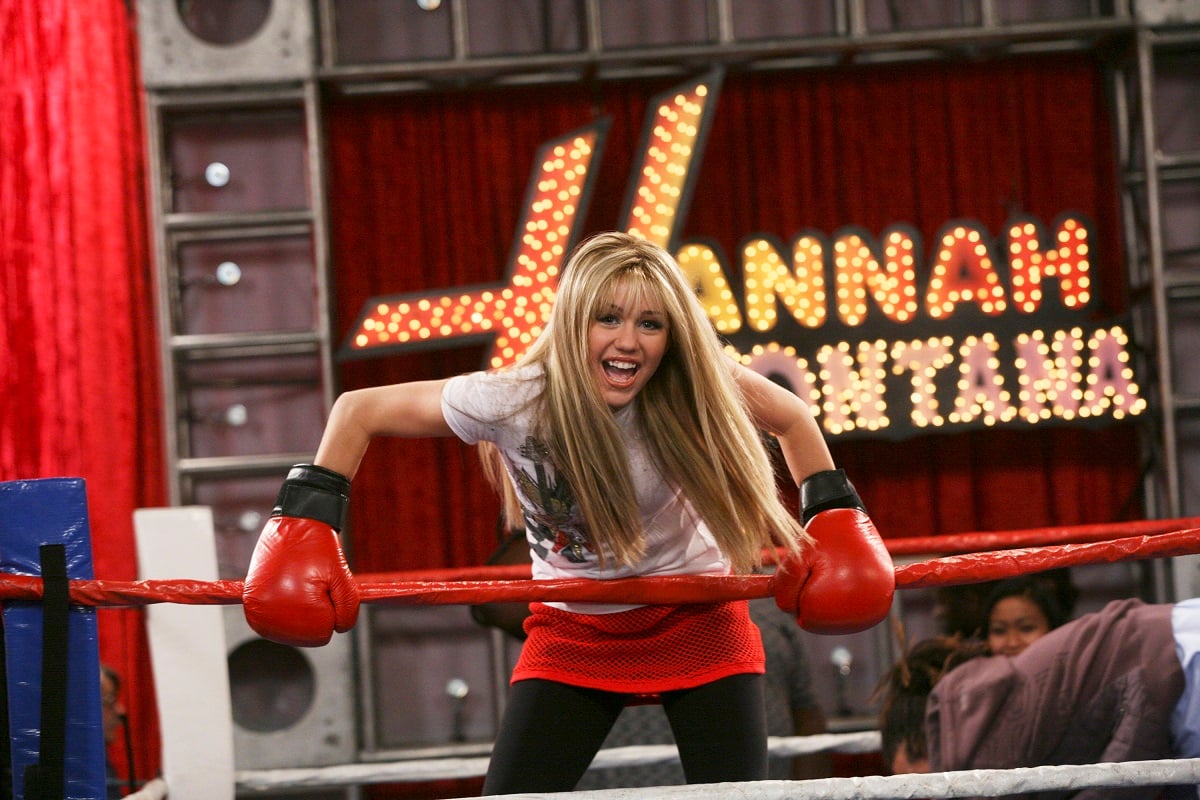 Miley Cyrus in a blonde wig wearing boxing gloves in front of a 'Hannah Montana' sign