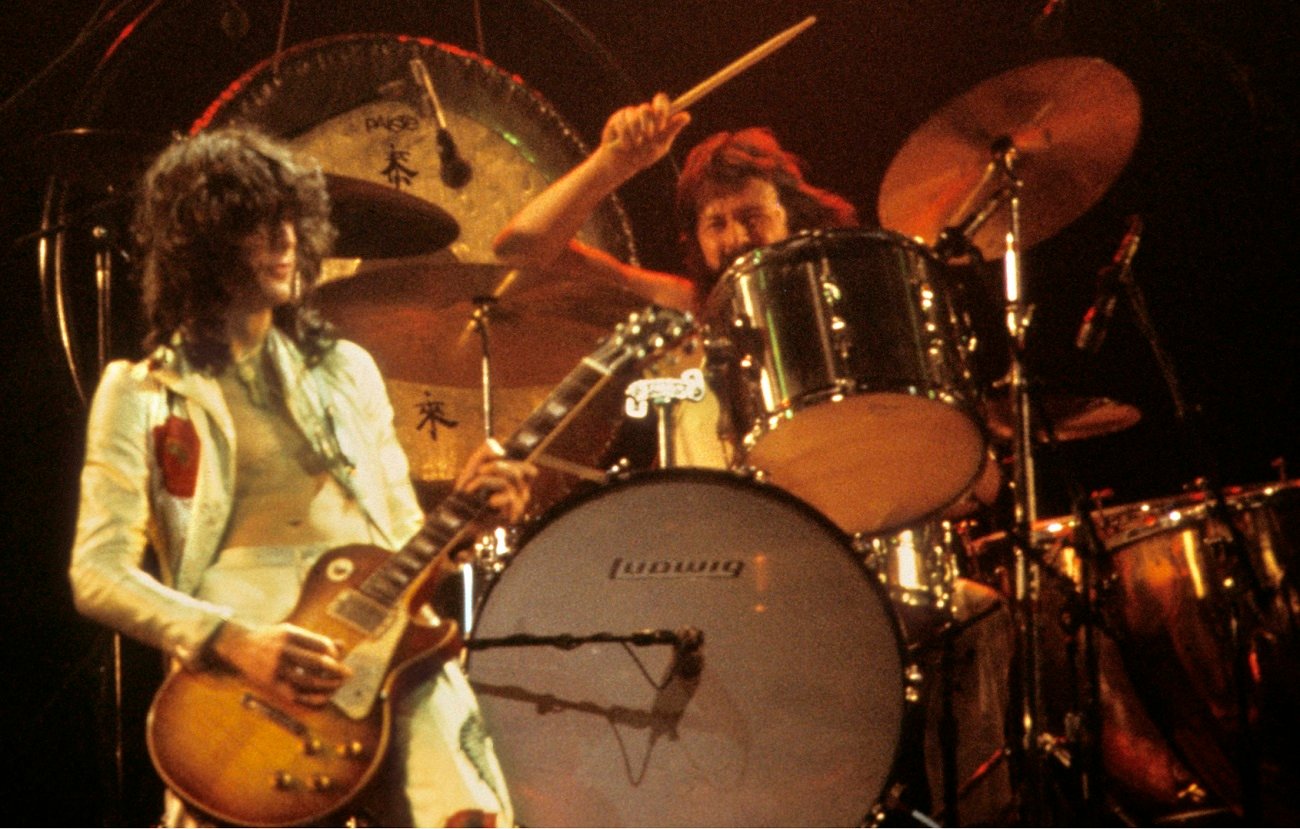 Jimmy Page plays guitar as John Bonham swings a drumstick behind him on stage in 1977