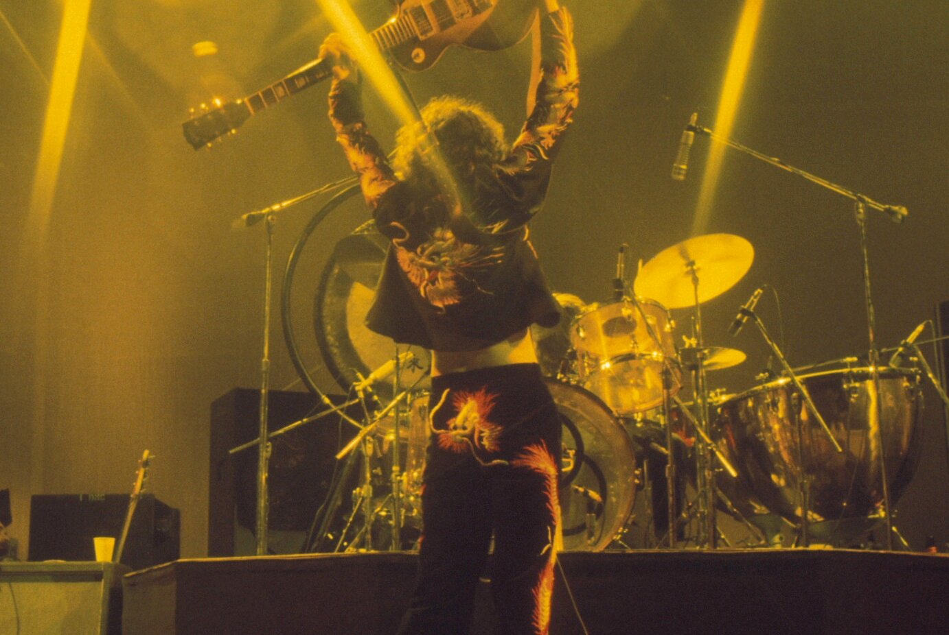 Jimmy Page, wearing a suit with colorful dragons sewn onto it, holds his guitar overhead on stage at Earl's Court, May 1975