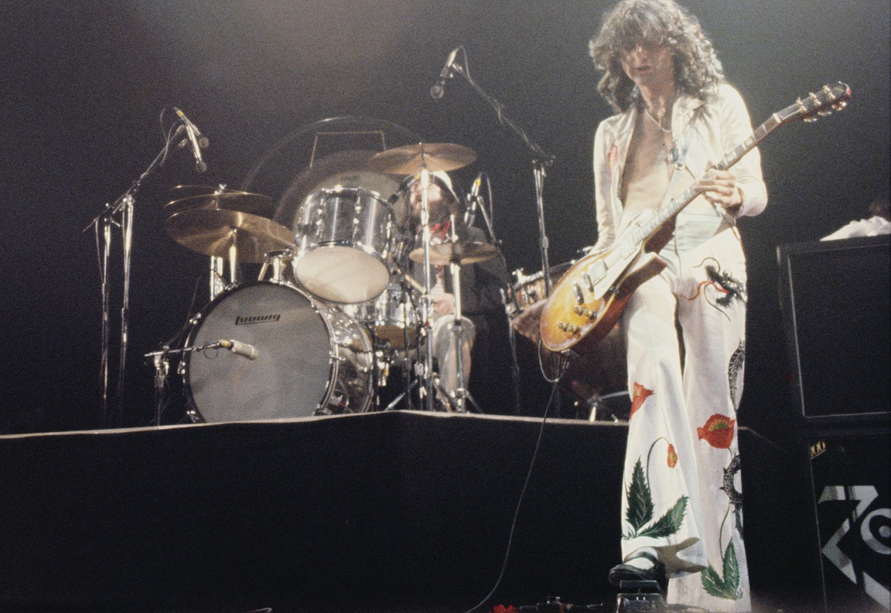 A view of John Bonham on drums with Jimmy Page playing guitar in a white suit with red poppies and other colorful objects on it in 1977