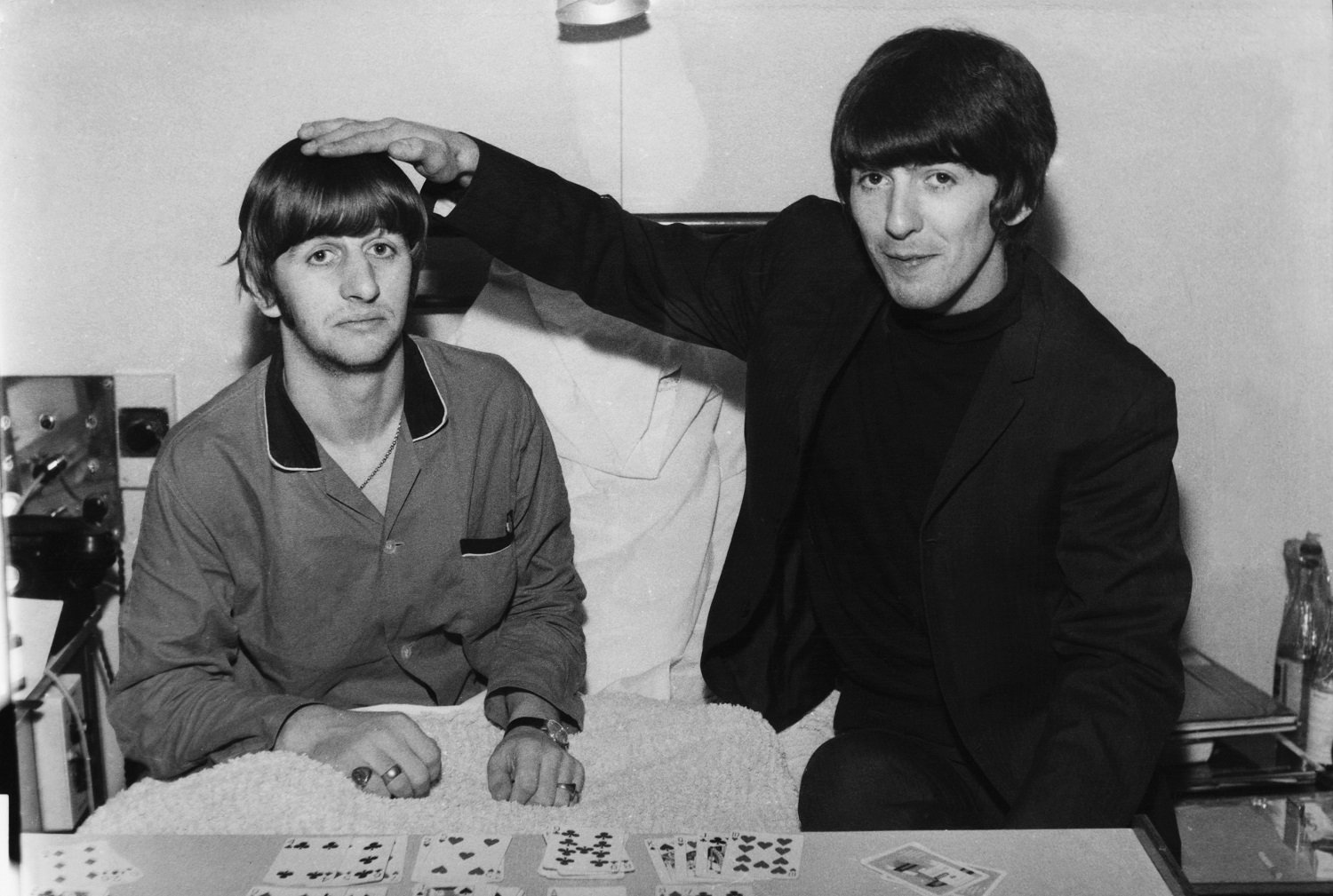 A smiling George Harrison holds his hand on top of Ringo Starr's head as the two sit in a hospital bed in 1964