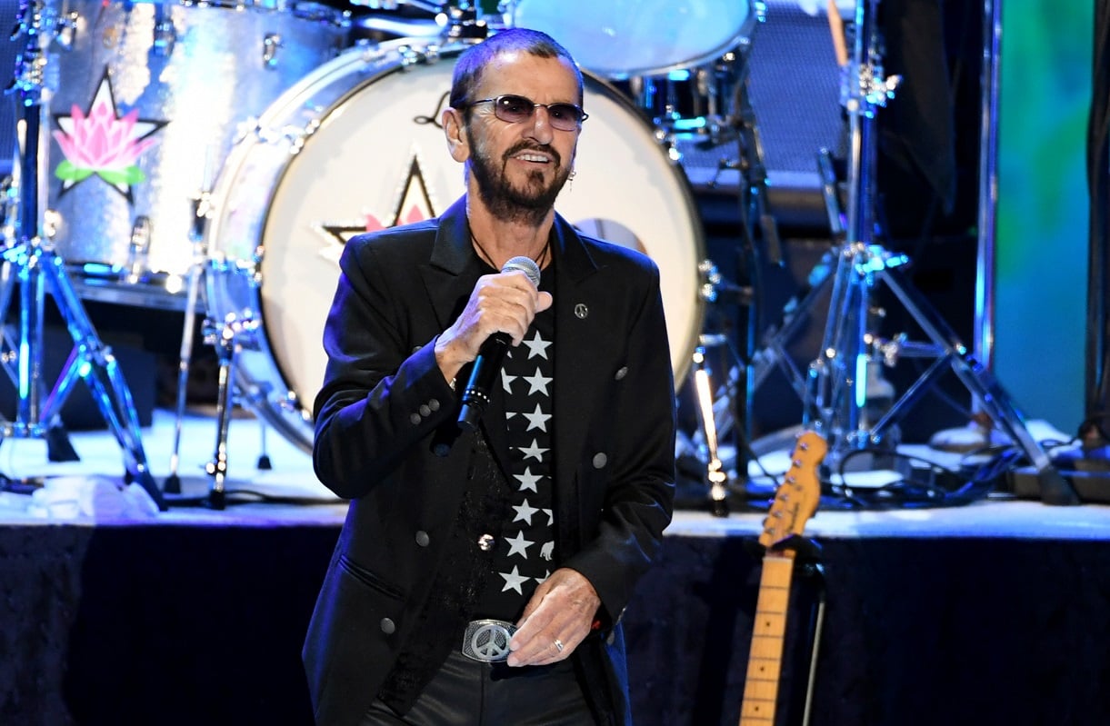 Ringo Starr performs on stage in 2019 wearing a black silver-buttoned jacket and dark-tinted glasses.
