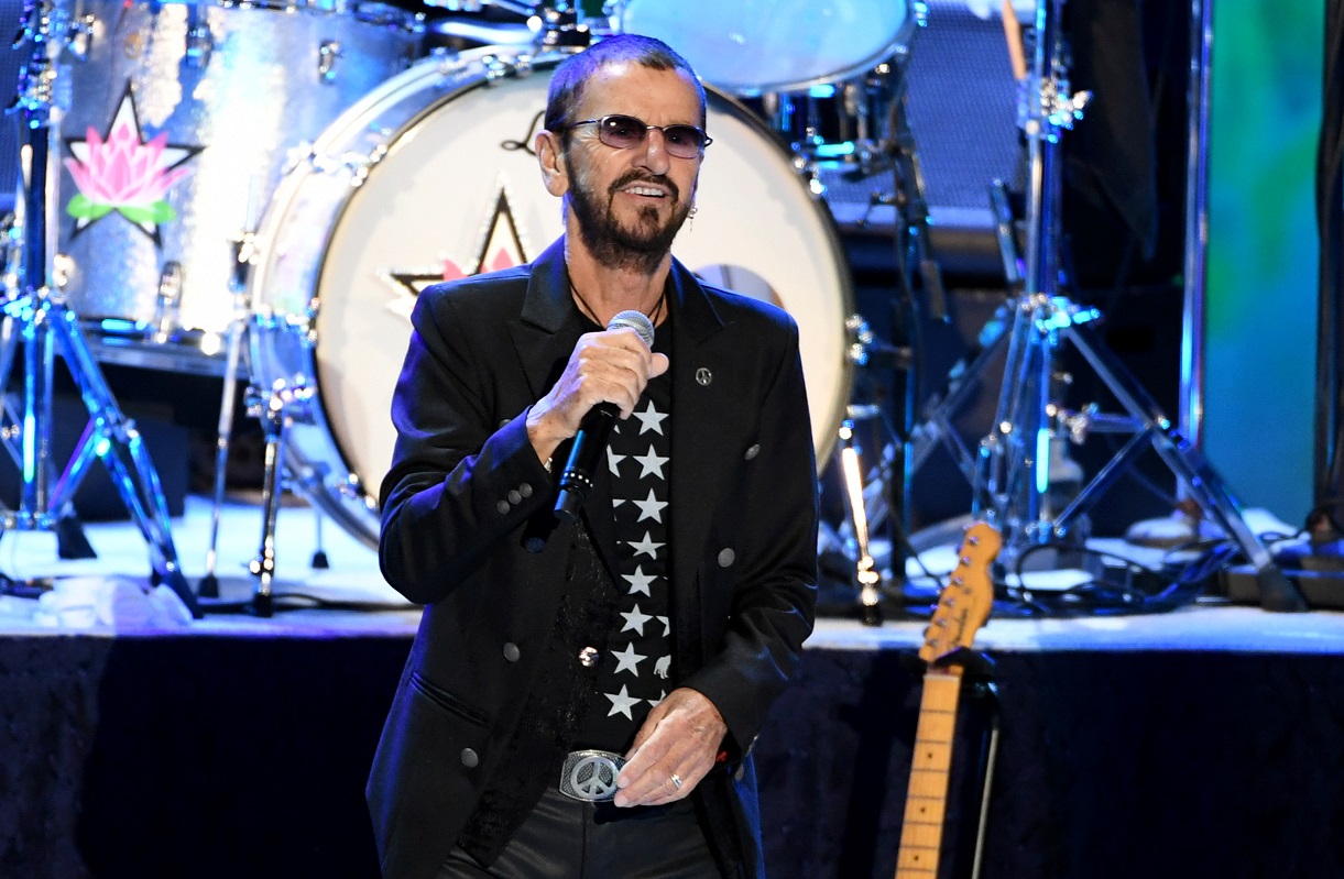 Ringo Starr performs on stage in 2019 wearing a black silver-buttoned jacket and dark-tinted glasses.