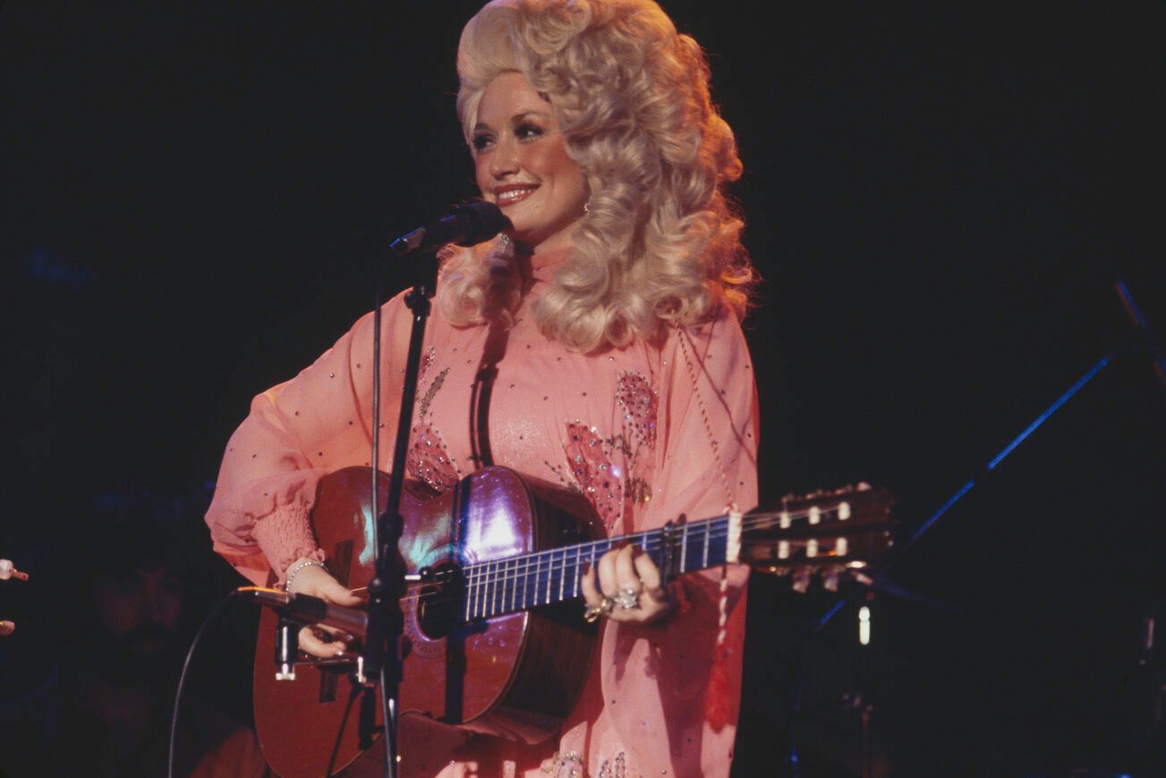 Dolly Parton performing on stage in New York in 1977. She's wearing a pink dress and big, blond, curly hair.