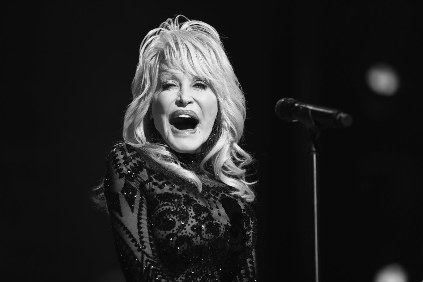 A black and white close-up of Dolly Parton singing into a microphone.