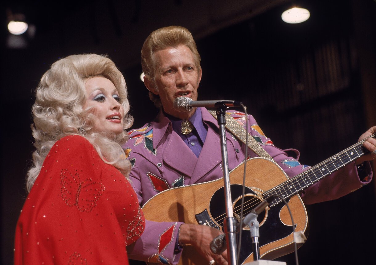 Dolly Parton and Porter Wagoner singing on stage in Nashville in 1978. Parton is in a bright red shirt with her hair in a beehive. Wagoner is in a purple jacket strumming a guitar.