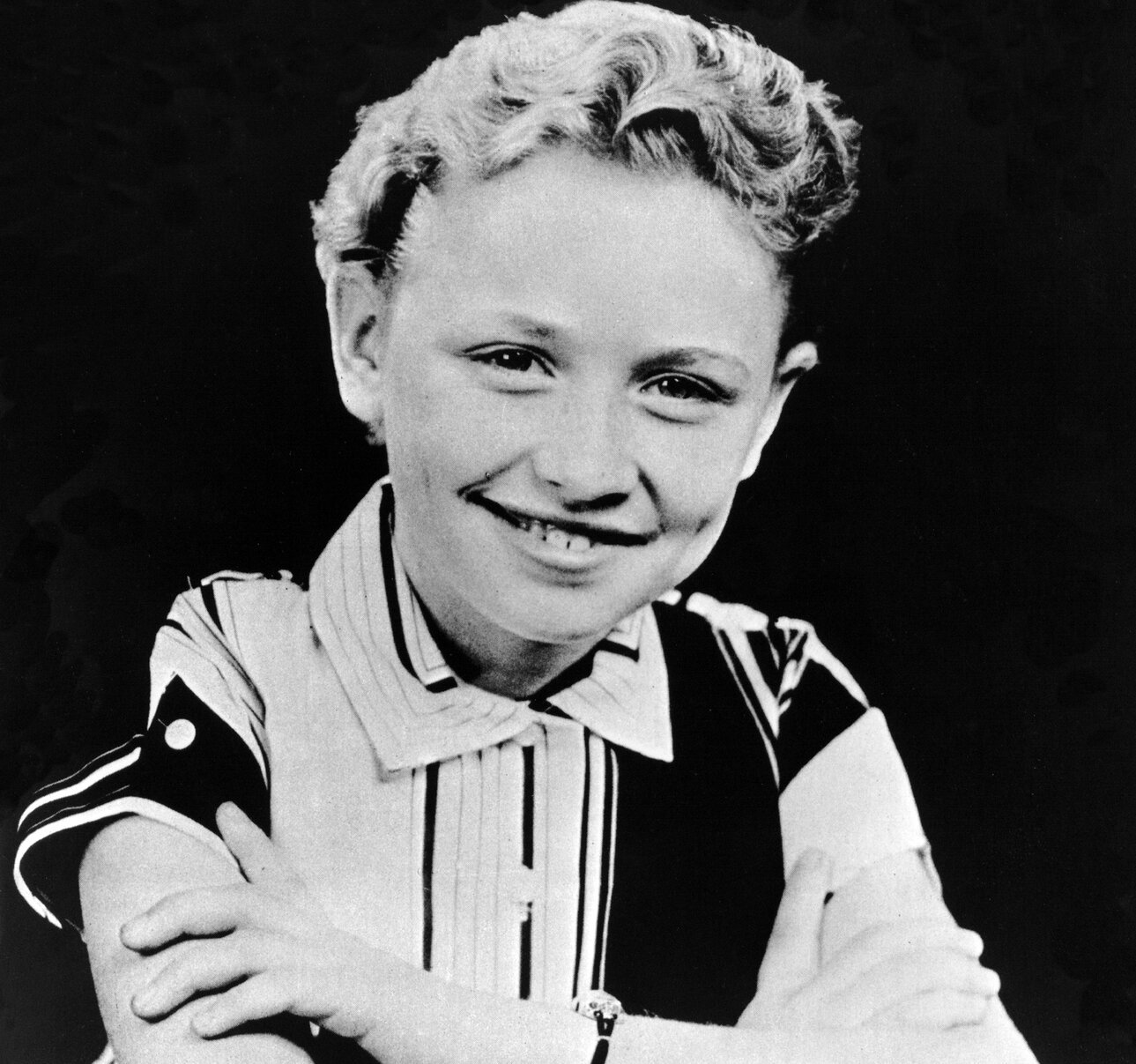 Dolly Parton poses for a portrait as a child in 1955. The photo is black and white. She wears short hair and a collared shirt.